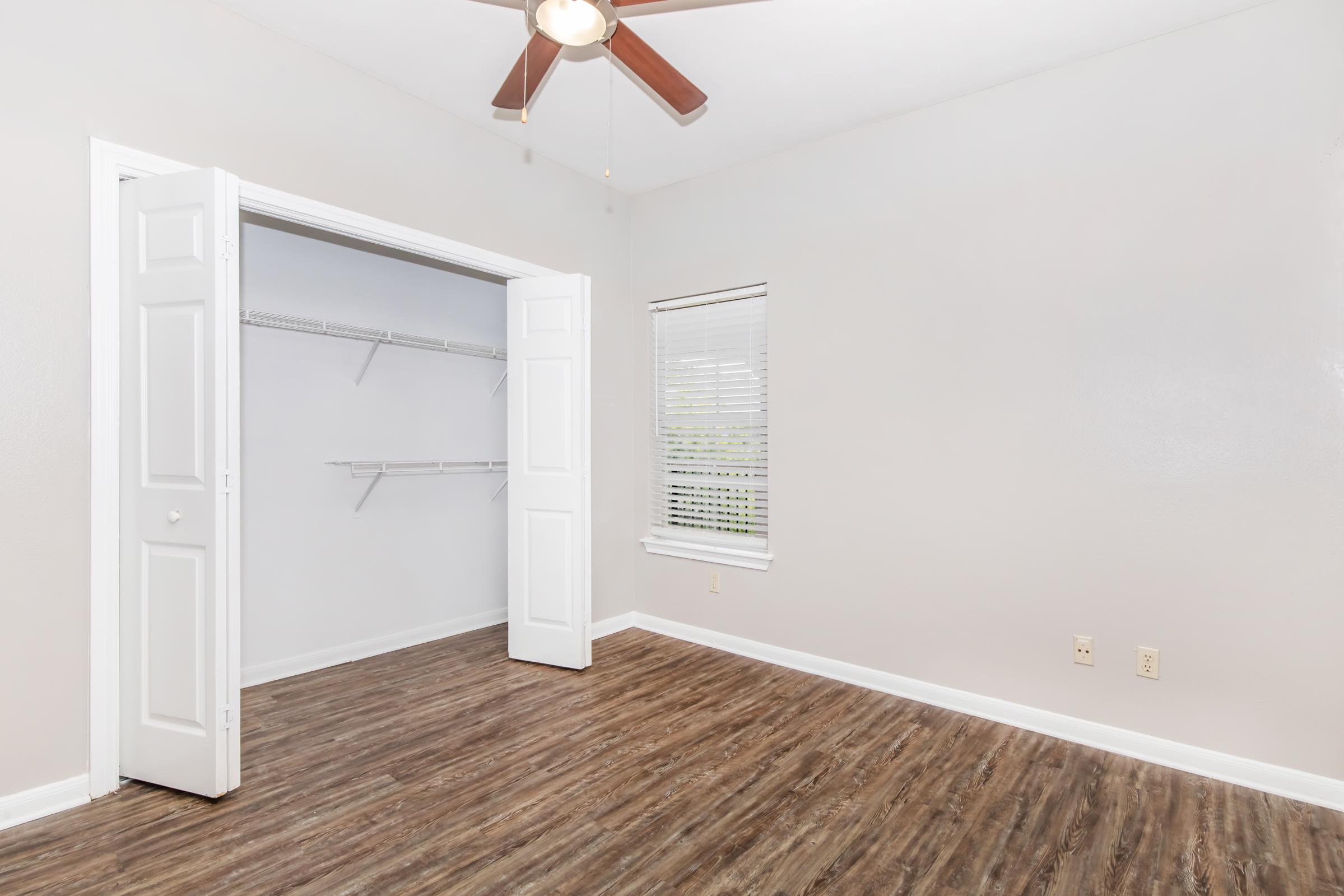 An empty room with light gray walls and a wooden floor. A ceiling fan is mounted on the ceiling, and there are two white doors leading to a closet with shelving. The window has blinds and allows natural light into the space.
