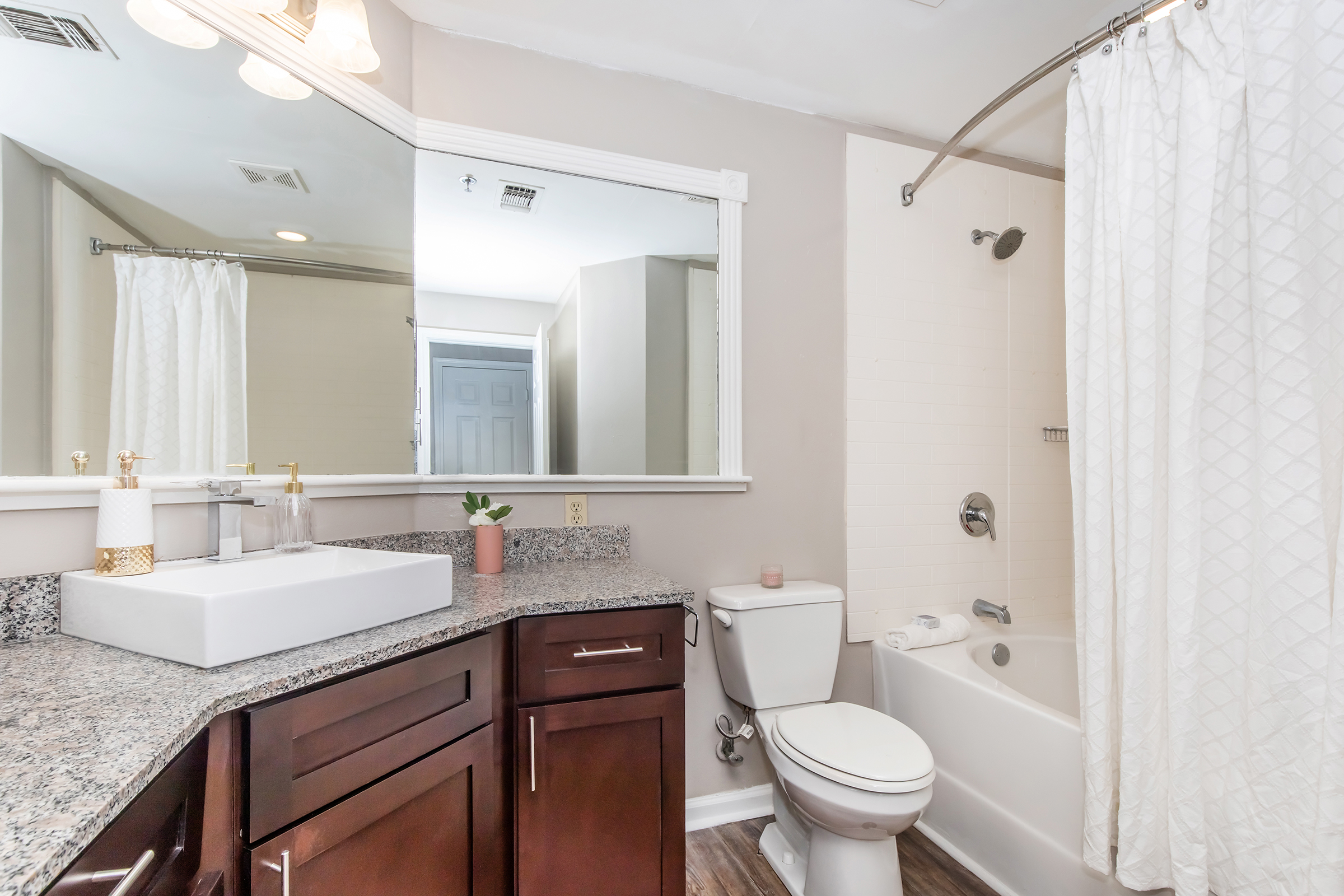 A modern bathroom featuring a white bathtub with a shower curtain, a sleek white sink on a dark wooden countertop, a mirror above the sink, and a small plant on the counter. The walls are painted a light gray, and there is a door visible in the background. Ample lighting from a ceiling fixture adds brightness to the space.