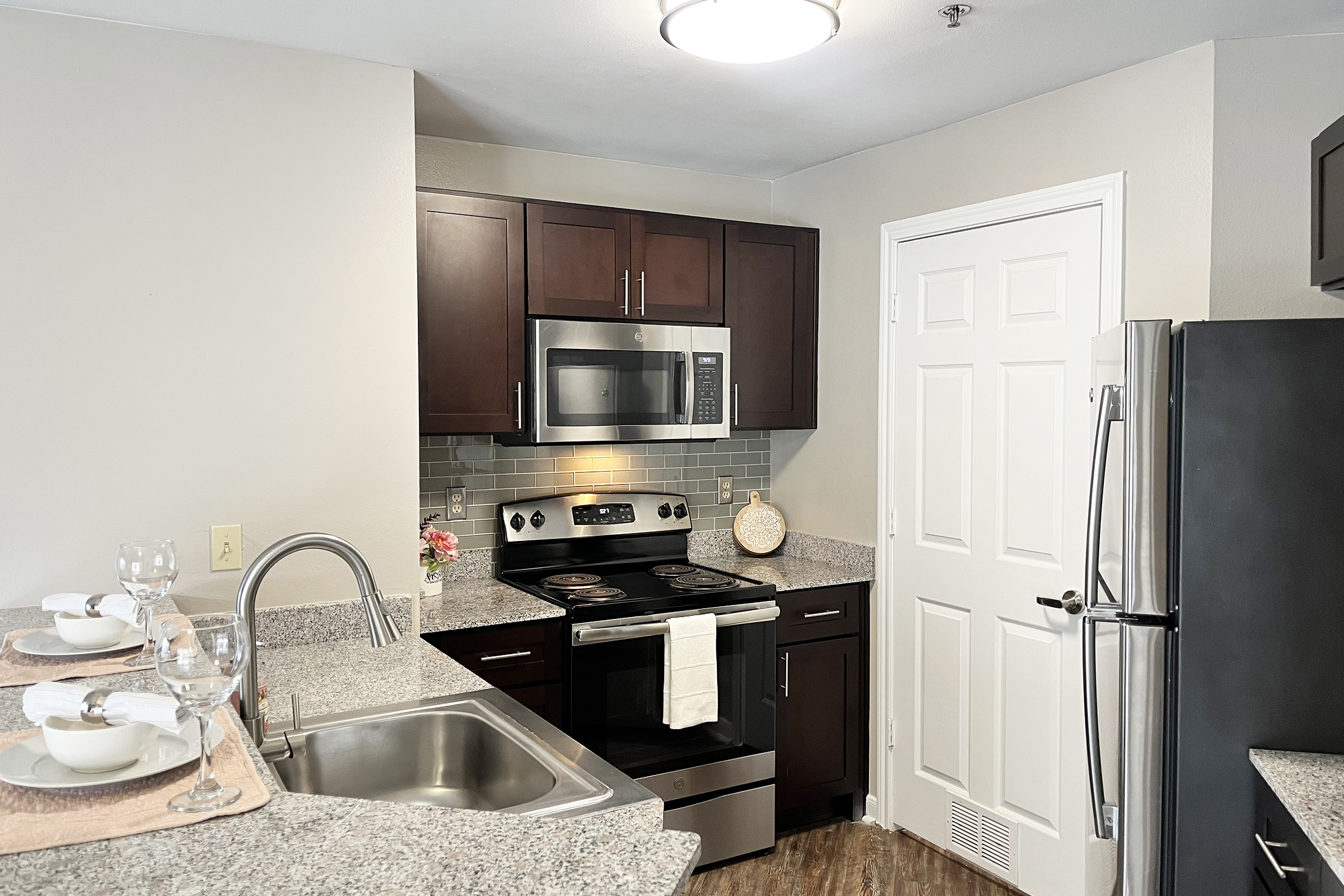 Modern kitchen featuring dark wood cabinets, stainless steel appliances, a granite countertop, and a sink. The kitchen includes a microwave, stove, and refrigerator, with decorative touches like a small flower arrangement. Natural light illuminates the space, creating a warm and inviting atmosphere.