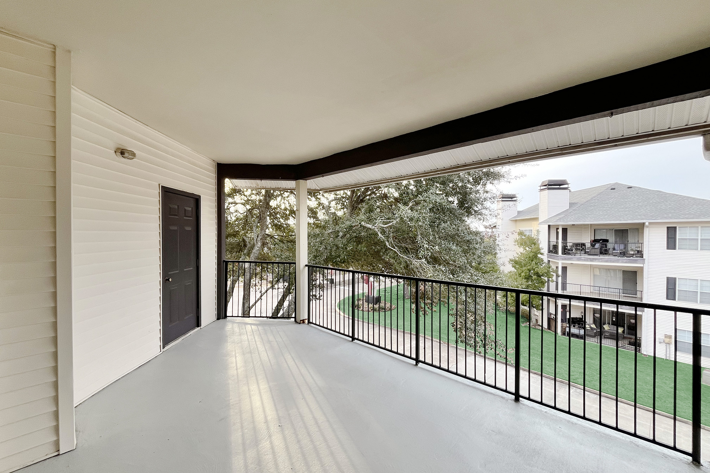 A spacious balcony with a black railing, overlooking a green lawn and surrounding buildings. The walls are white with a solid black door. Natural light casts shadows on the floor, creating a bright and airy atmosphere.