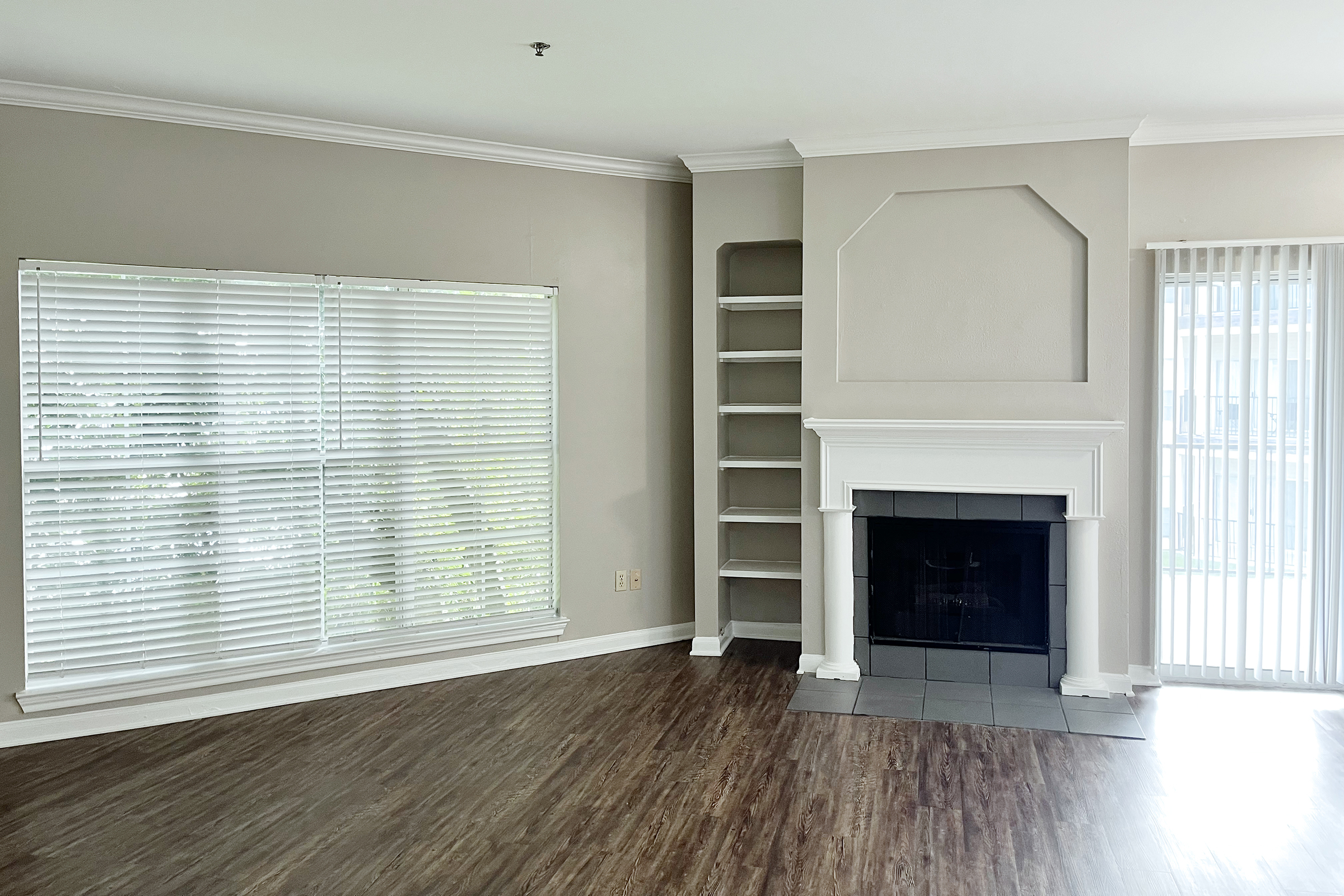 A spacious living room featuring hardwood flooring, a large window with white blinds, built-in shelves, and a white fireplace with a simple design. Natural light fills the room, creating a warm atmosphere.