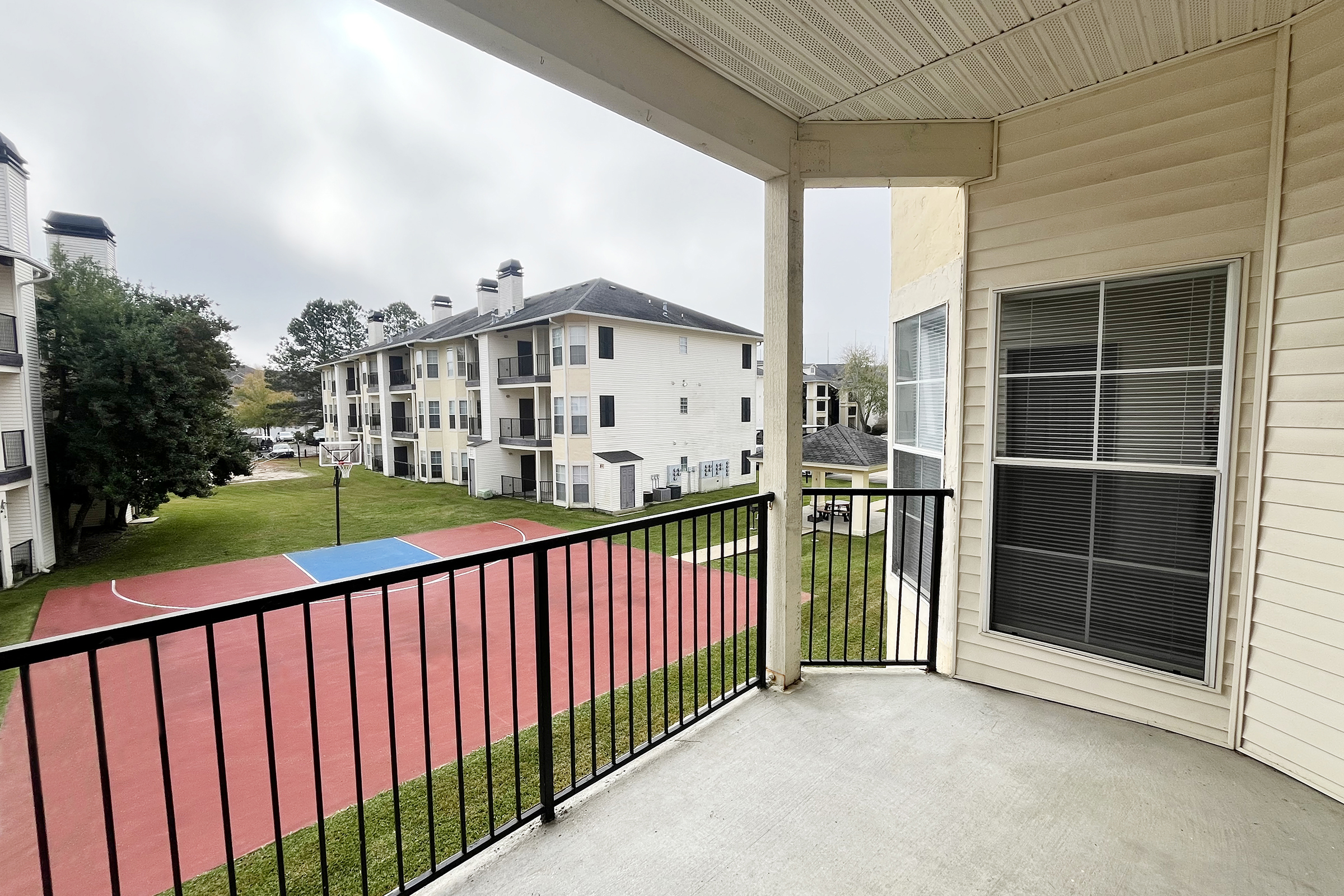 View from a balcony overlooking a basketball court and landscaped area of an apartment complex. The scene features a cloudy sky, with several buildings visible in the background. The court is painted in blue and red, surrounded by green grass.