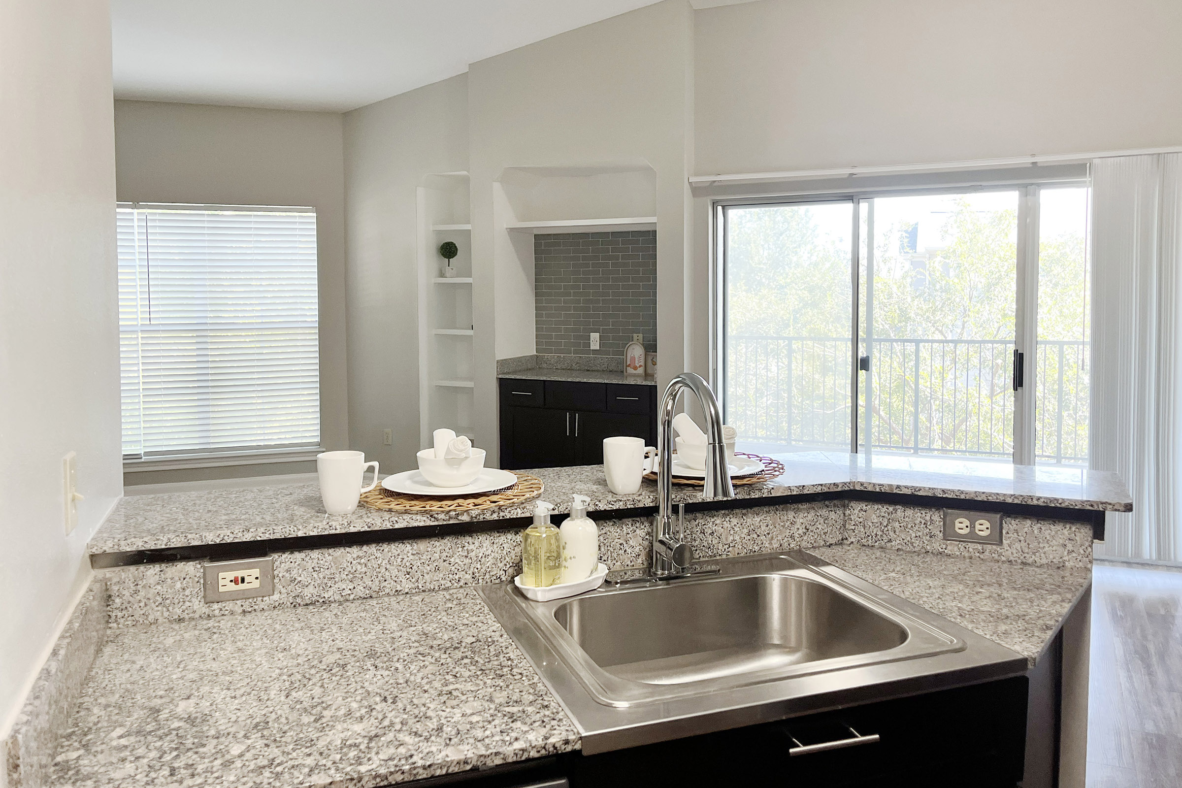 A modern kitchen featuring a granite countertop, stainless steel sink, and dishes arranged on a placemat. In the background, a living area with a large window allows natural light, showcasing a view of greenery outside. Shelving units are visible, complementing the contemporary design.