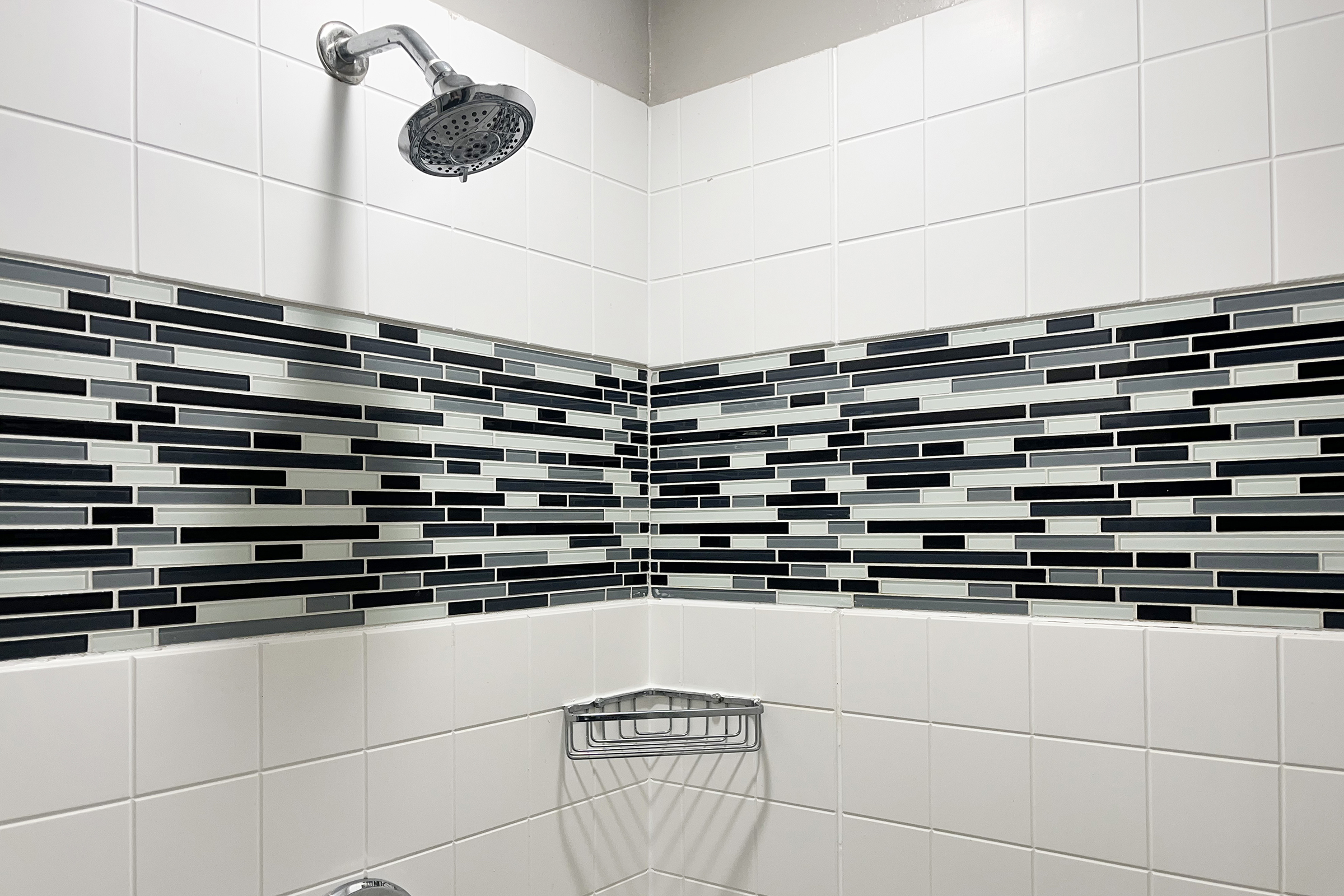 A modern shower corner featuring a chrome showerhead and a mix of black, white, and gray rectangular tiles arranged in a contemporary pattern. The walls are primarily white with a decorative border of stylish tiles, enhancing the bathroom's aesthetic.