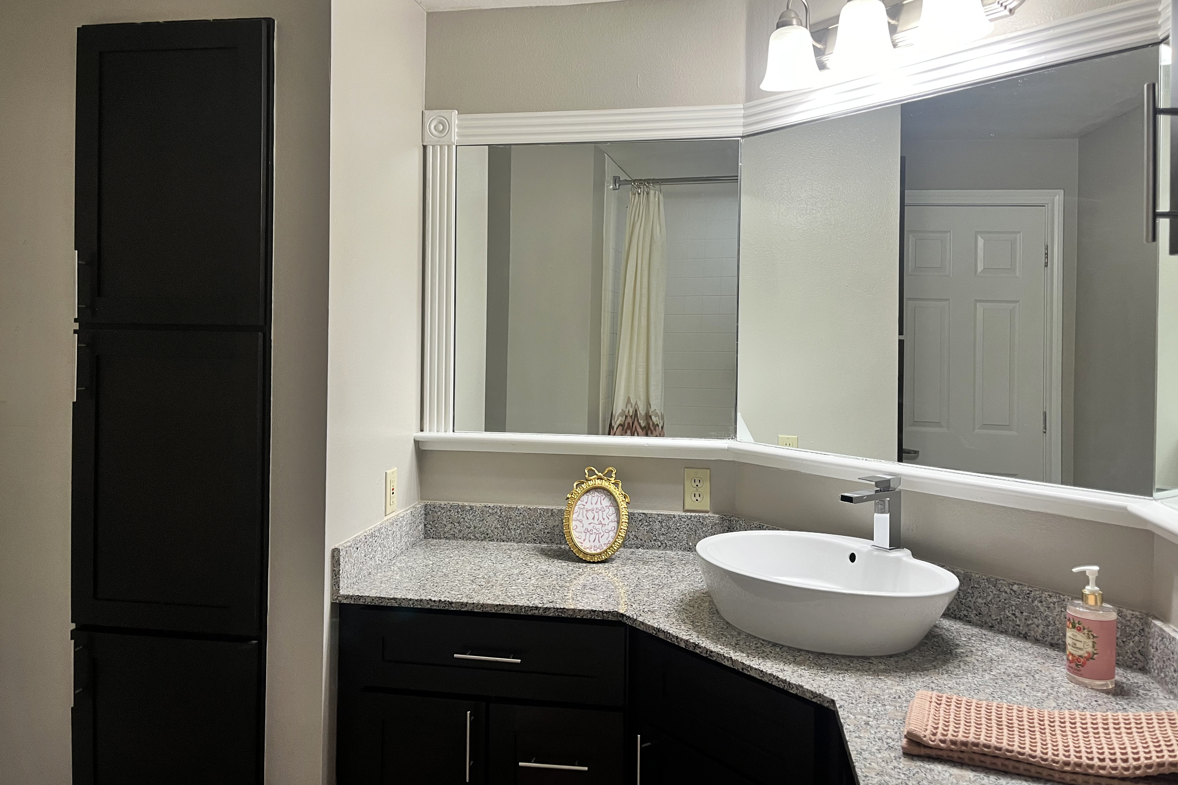 A modern bathroom featuring a granite countertop with a white vessel sink, a stylish faucet, and a decorative mirror. The wall is mostly light-colored, contrasting with the dark cabinetry. There's a shower visible in the background and a cute framed decorative piece on the counter next to a bottle of hand soap.