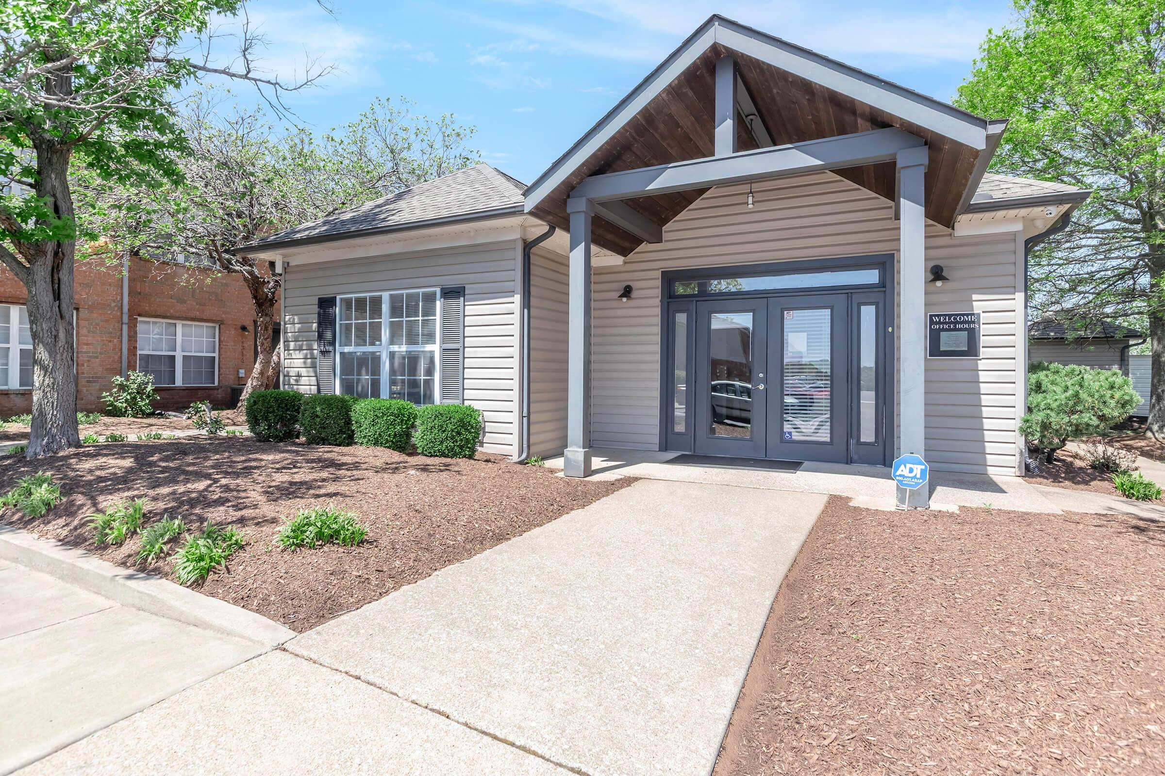 A modern single-story building with a peaked roof and large front entrance. The exterior features light gray siding and a welcoming pathway leading to the door. Lush green shrubs and freshly mulched flower beds enhance the landscape, with trees visible in the background and a security sign near the entrance.