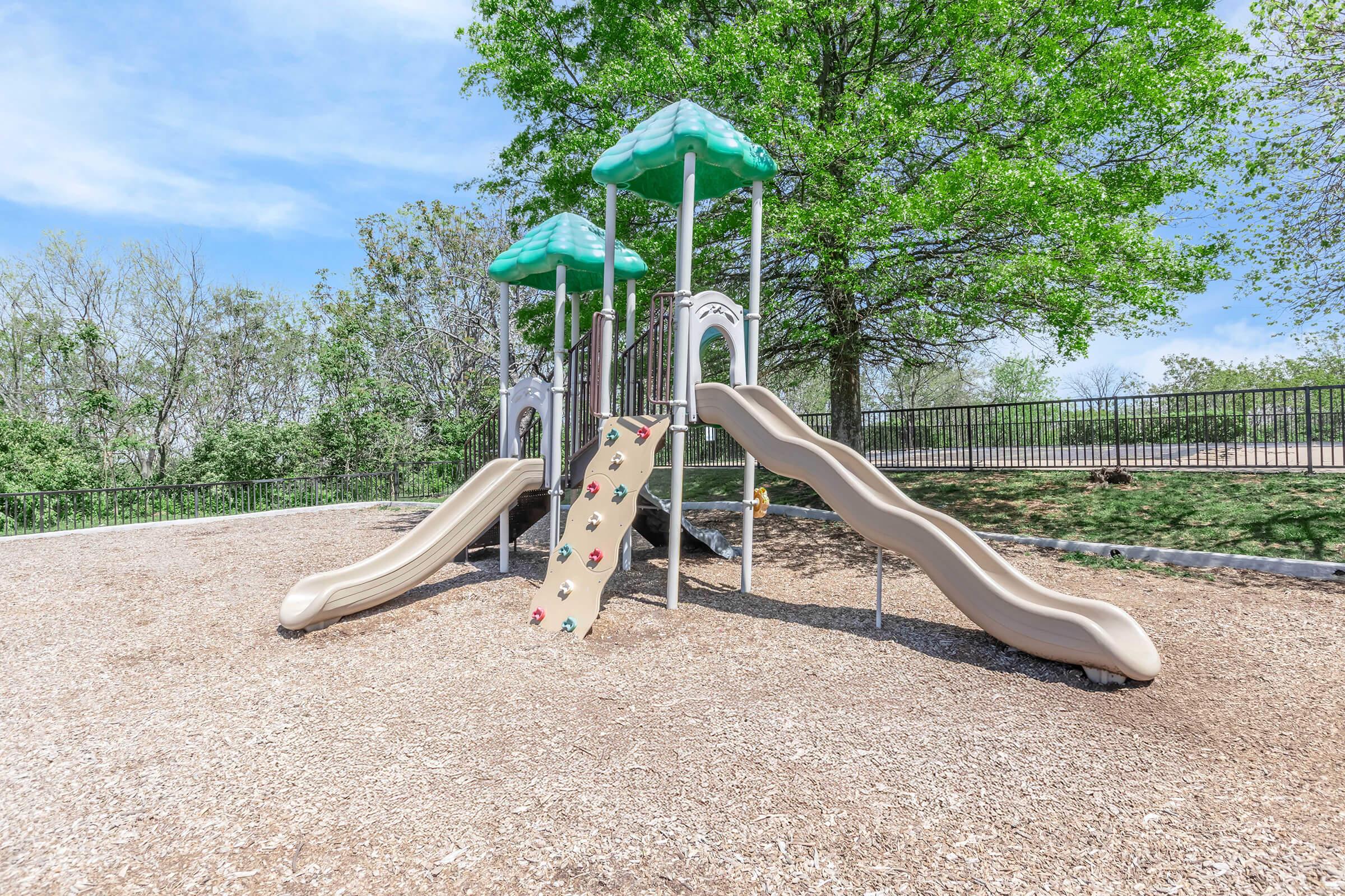 Playground equipment featuring two green-roofed slides set against a backdrop of trees. The ground is covered with wood chips, and there is a grassy area in the distance. The playground is well-maintained, providing a safe environment for children to play.