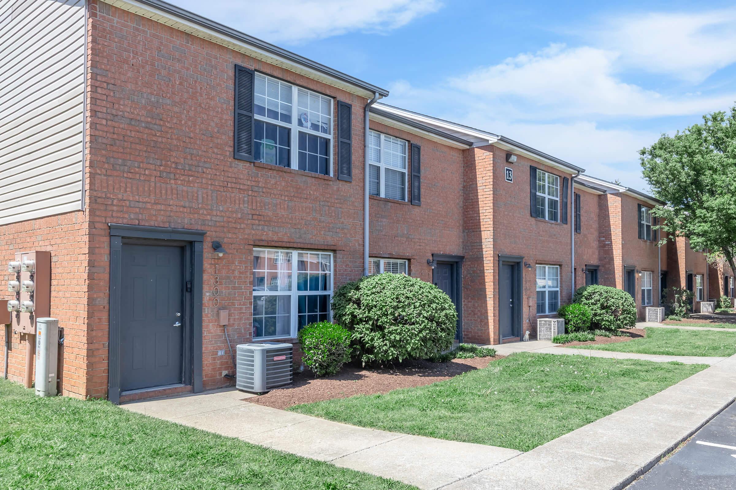 A row of red brick apartment buildings with large windows and air conditioning units. Each unit has a front door and is flanked by neatly trimmed bushes. The area is well-maintained with green grass and a clear blue sky overhead.