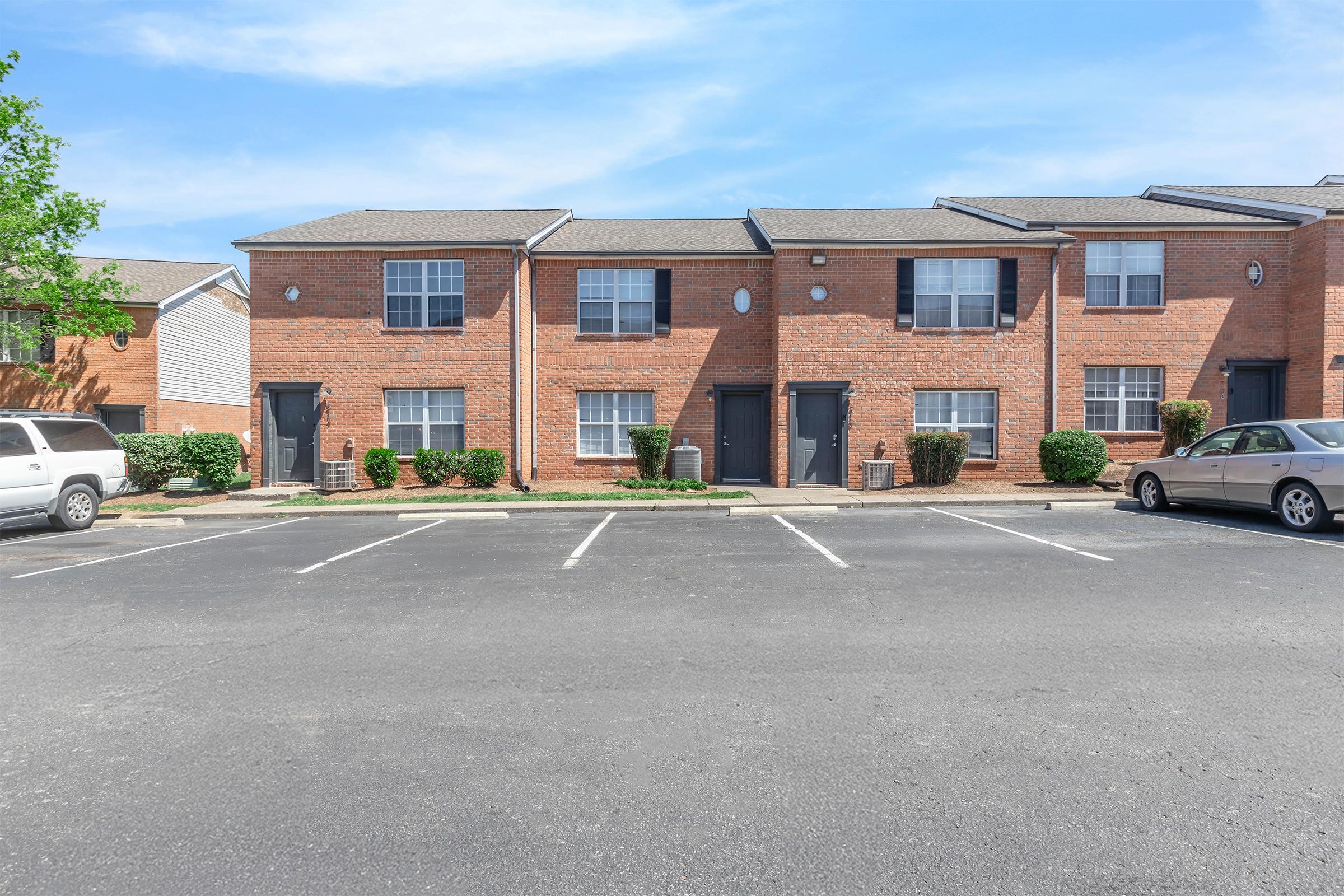 A row of brick apartment buildings with multiple entrances, situated in a parking lot. The scene features several parked cars and landscaped bushes, under a clear blue sky. The buildings have green front doors and multiple windows, creating a residential neighborhood atmosphere.