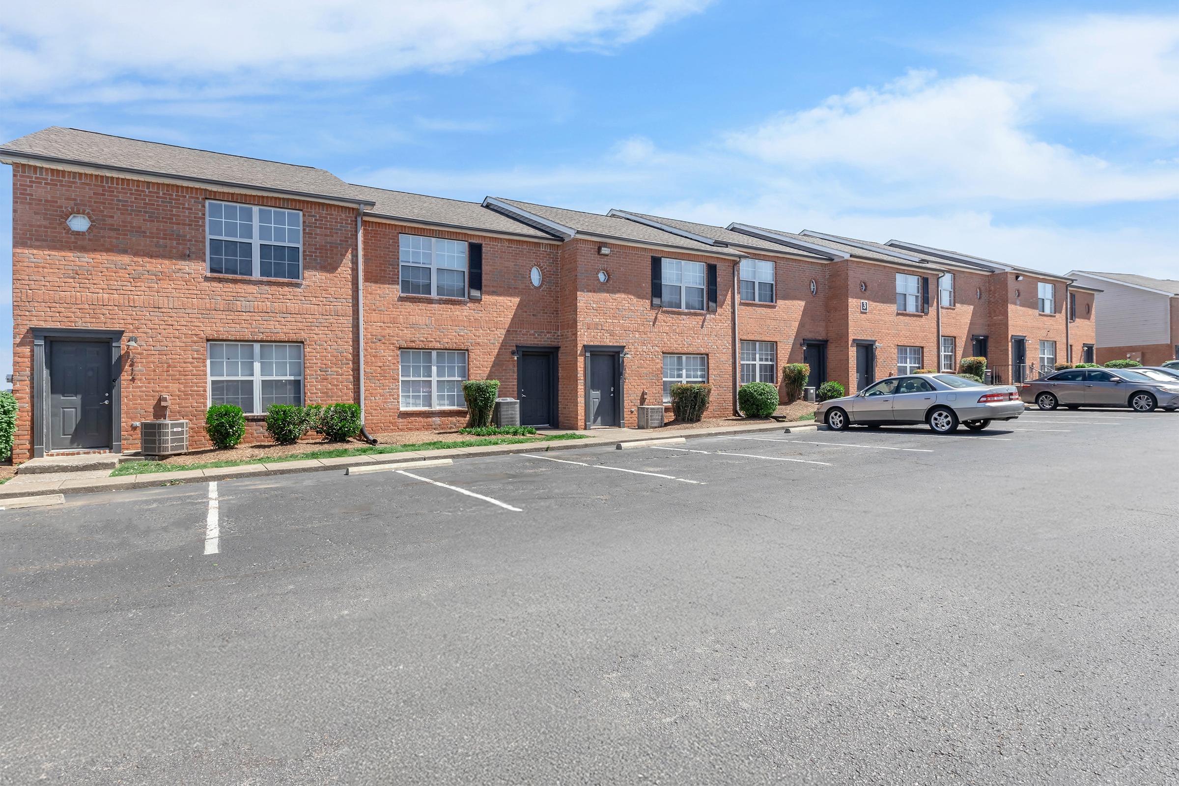 Row of brick apartment buildings with gray roofs, featuring windows and parked cars in front. Neatly trimmed bushes line the walkway, and a clear blue sky is visible above.