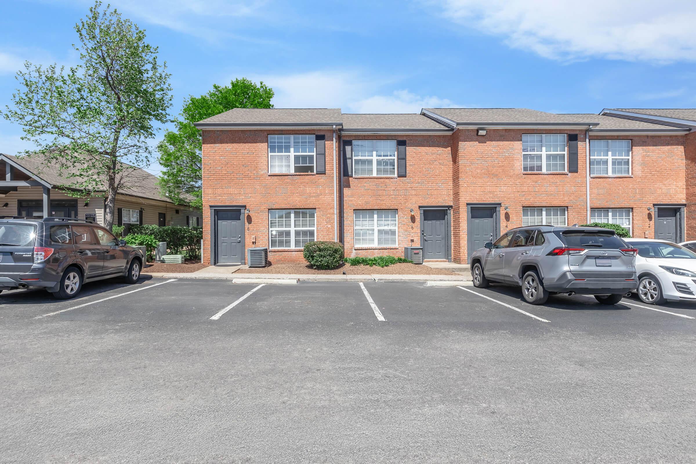 Two-story brick apartments with several parked cars in the foreground. Each unit has windows with white frames and a small bush in front. The sky is clear with a few clouds, and there are trees nearby, adding to the residential atmosphere.
