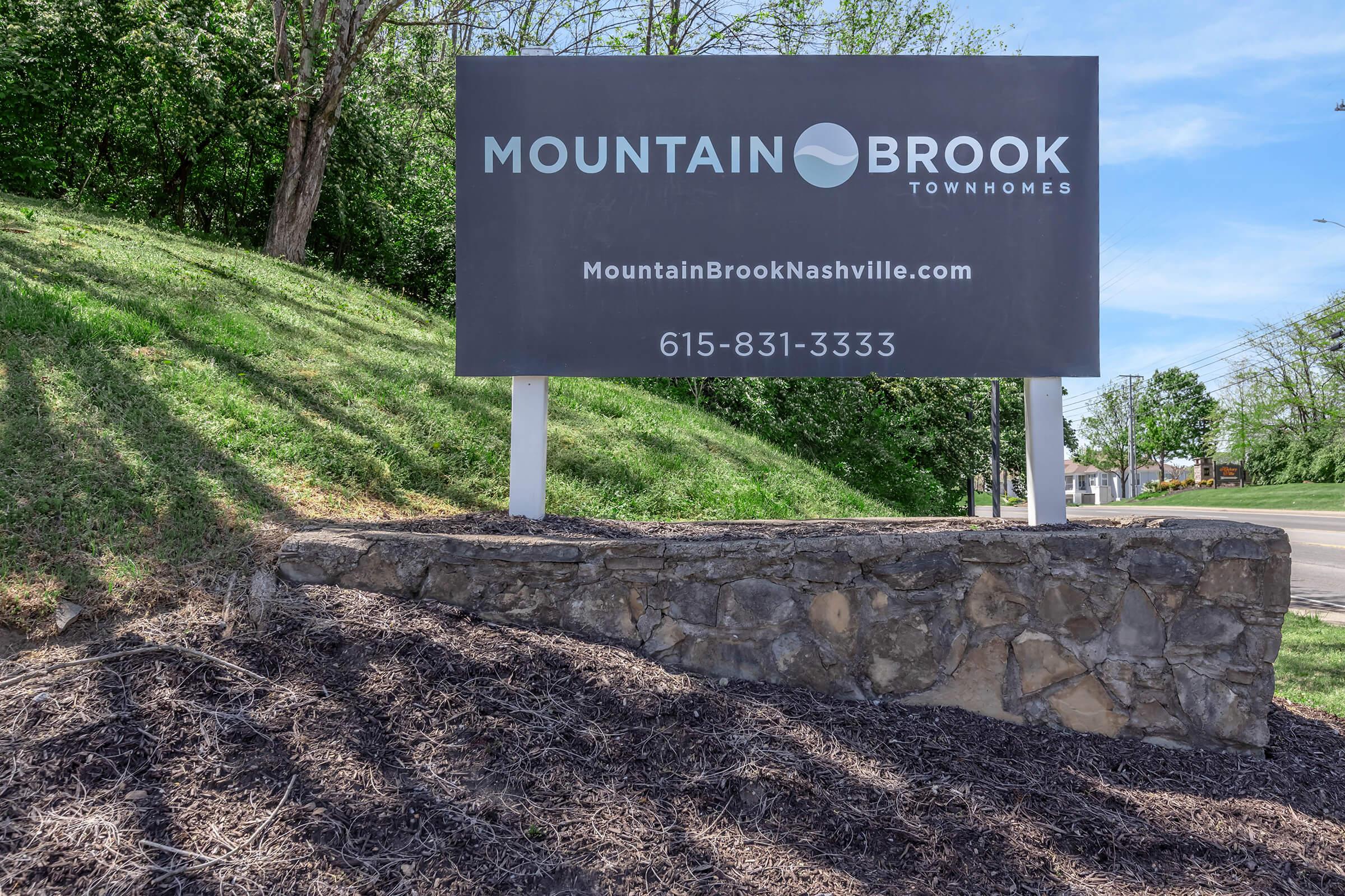 Sign for Mountain Brook Townhomes featuring the name "Mountain Brook" prominently displayed. It includes a website address, "MountainBrookNashville.com," and a phone number "615-831-3333." The sign is set against a green, grassy background with a stone base.