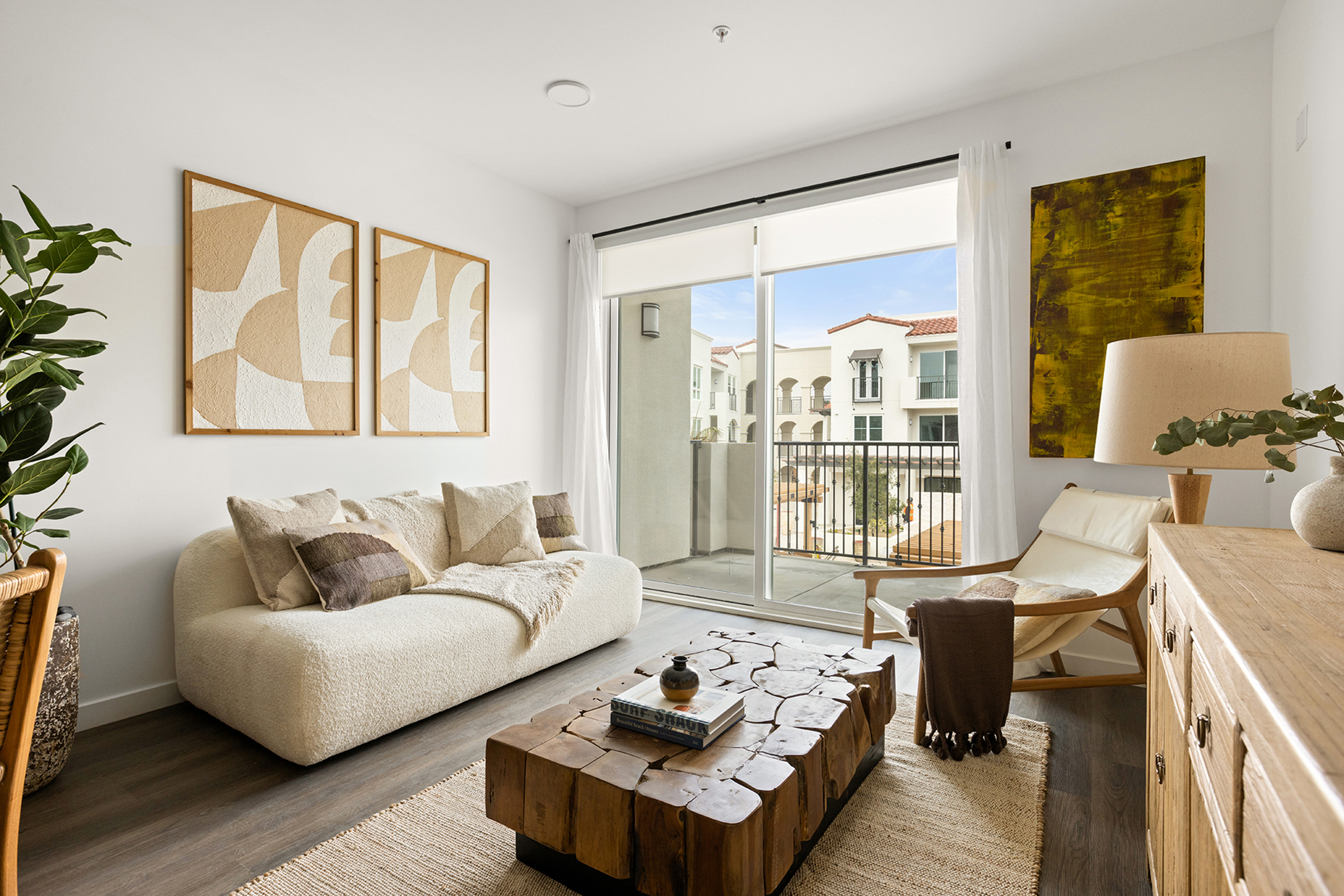 A modern living room featuring a cozy white sofa adorned with decorative pillows, a wooden coffee table made of logs, and a light-colored armchair. Large windows provide natural light, revealing a balcony view. The walls are decorated with abstract artwork, and potted plants add a touch of greenery to the space.