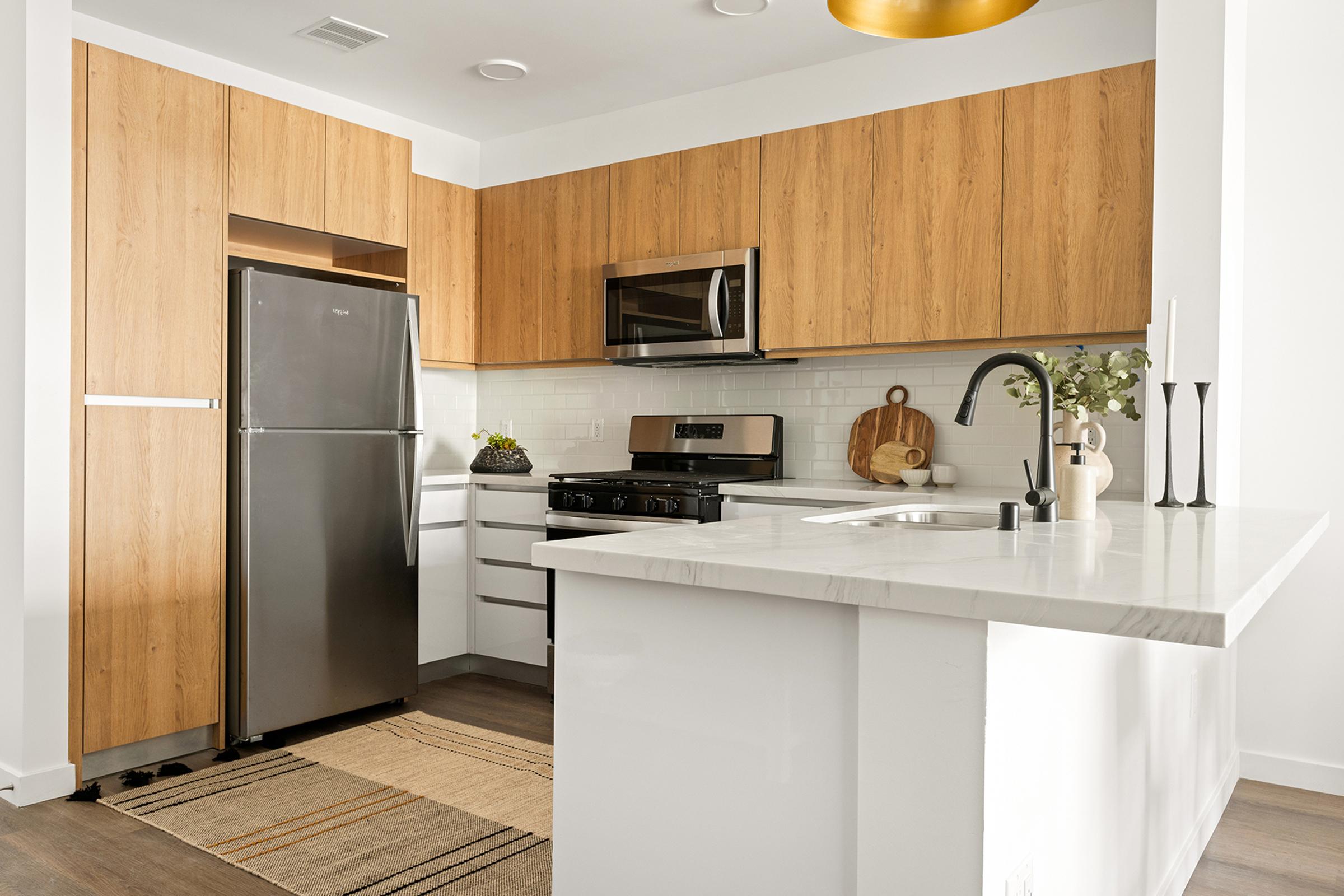 Modern kitchen featuring wooden cabinetry, a stainless steel refrigerator, an oven with a microwave above, and a marble countertop. The space includes a sink and decorative items like a cutting board and plants, with a stylish light fixture above.