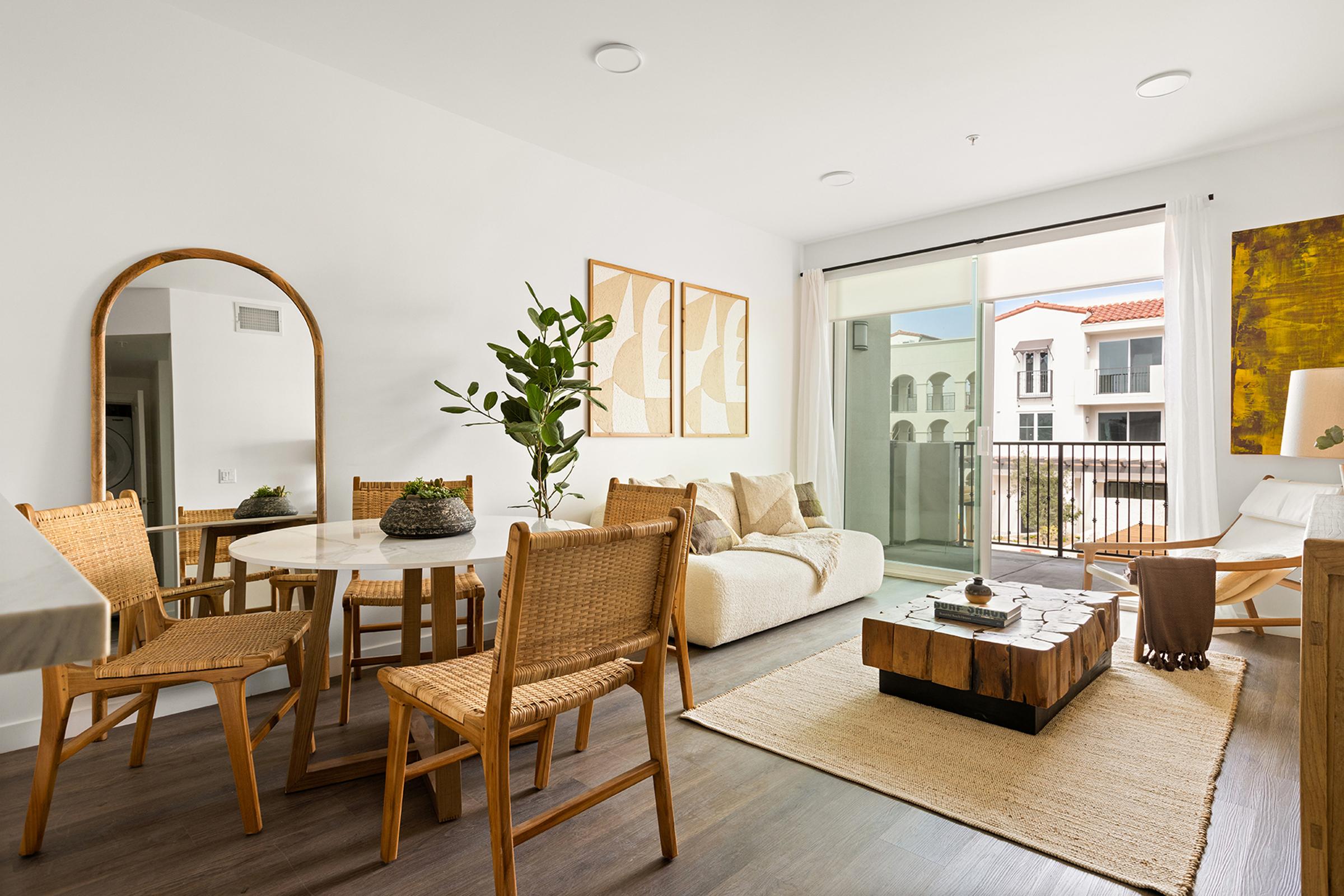 A modern, bright living room featuring a round dining table with four wicker chairs, a cozy sofa, and a wooden coffee table. Large windows provide natural light, facing a balcony. Decorative plants and art pieces add a touch of style, while a woven rug enhances the warm ambiance.