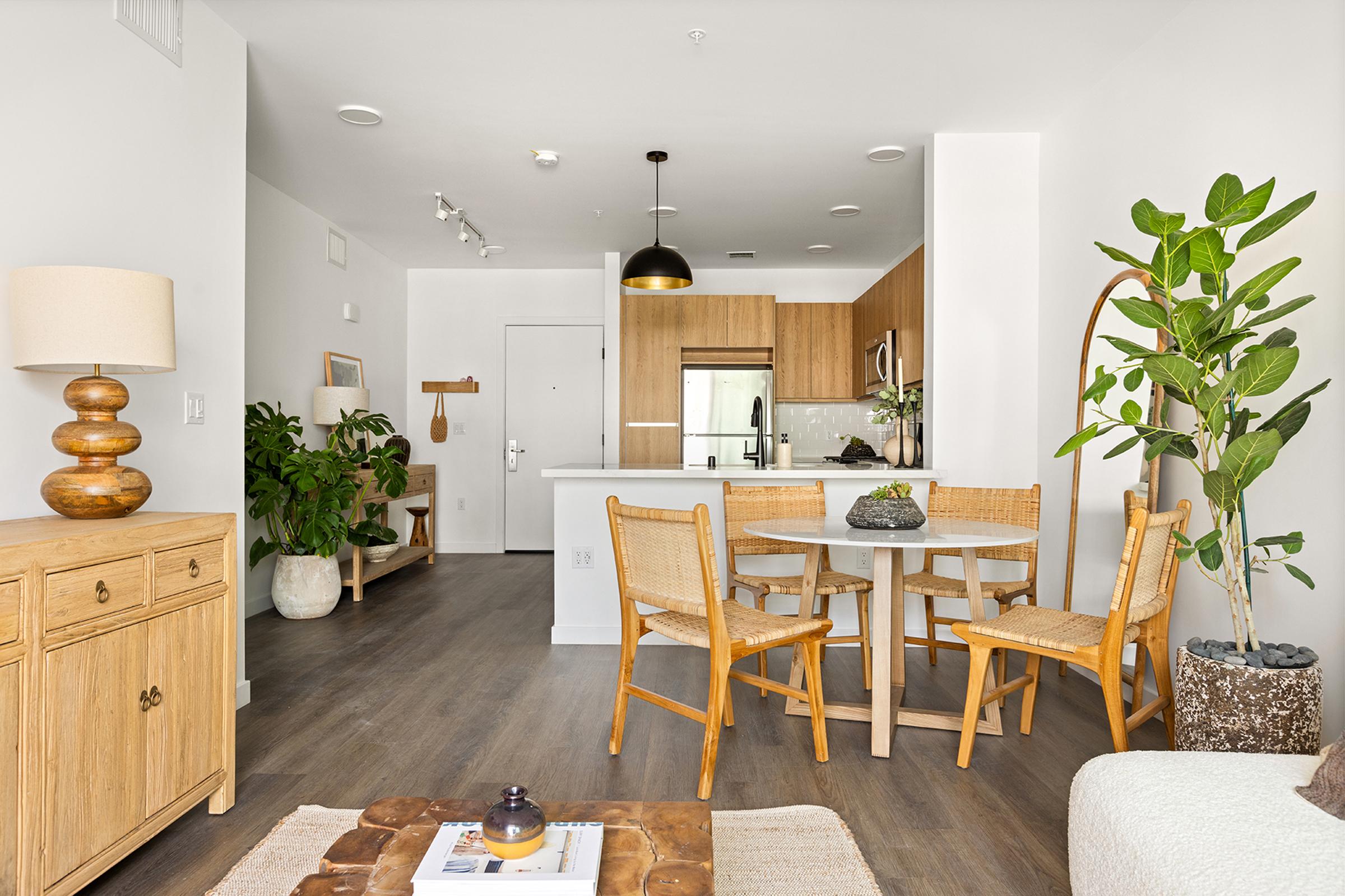 A modern, bright open-concept living space featuring a small dining area with a round table and four chairs, a beige sofa, a wooden sideboard, and decorative plants. The kitchen area is visible in the background, showcasing wood cabinetry and stainless steel appliances, with light walls and hardwood floors.