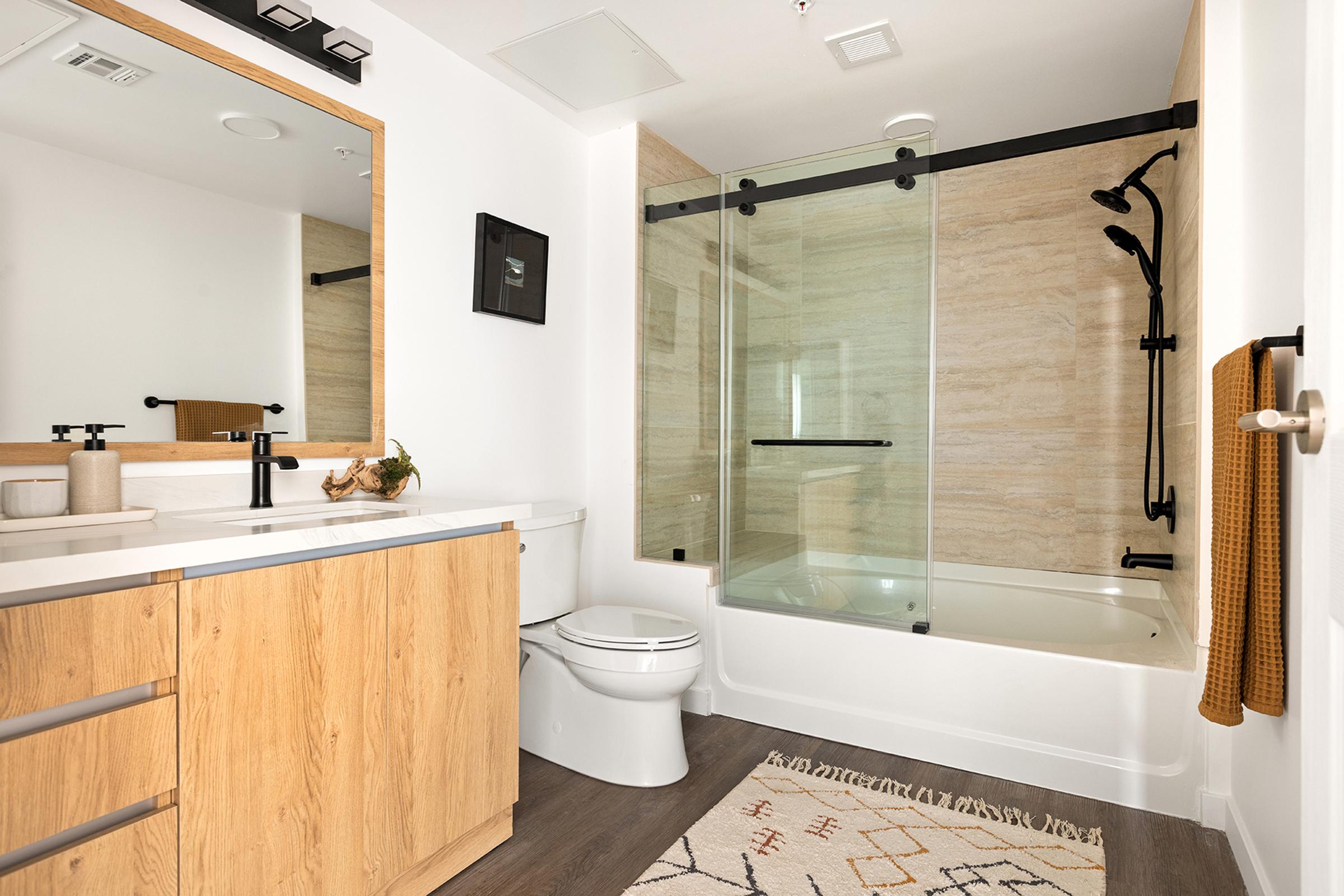 Modern bathroom featuring a sleek design with a large mirror, wooden cabinetry, and a bathtub with a glass shower enclosure. A towel hangs on the side, and decorative items are placed on the counter. Warm lighting highlights the neutral color palette and textured wall.