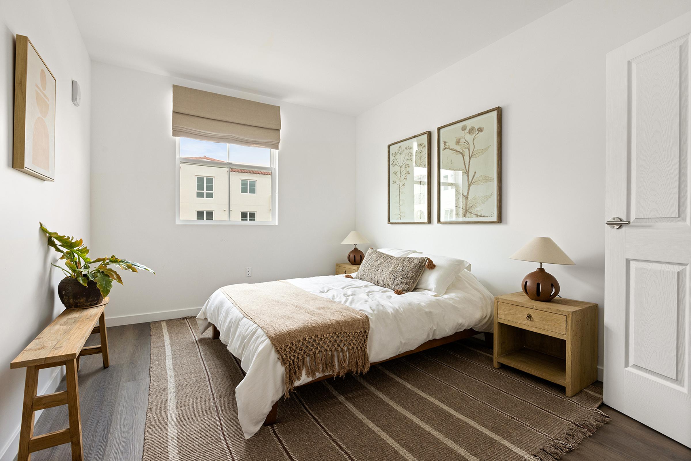 A bright and minimalistic bedroom featuring a queen-sized bed with white linens, a decorative throw blanket, and two bedside tables with lamps. A wooden bench is positioned against the wall, and botanical art prints are framed and hung above the bed. Natural light streams in through a window with a light shade.