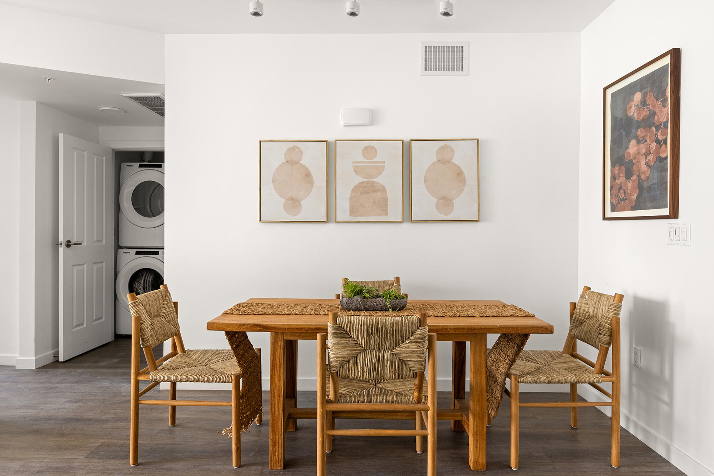 A modern dining area featuring a wooden table surrounded by four chairs with woven seats. On the wall, there are three minimalist framed artworks. A doorway leads to a laundry area with a washer and dryer visible. The walls are white, and the flooring is a dark wood, creating a warm and inviting atmosphere.