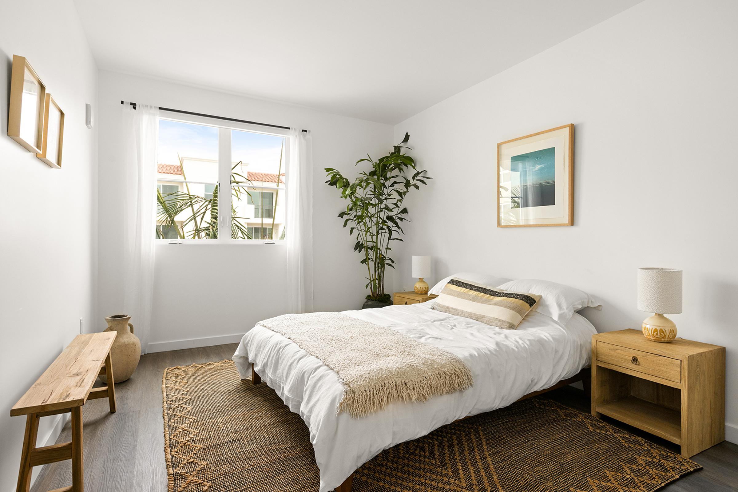 A cozy, modern bedroom featuring a queen-sized bed with white bedding, a textured throw, and decorative pillows. There are two nightstands with lamps, a wooden bench, and a decorative pot. A large window lets in natural light, complemented by indoor plants and minimal decor for a serene atmosphere.