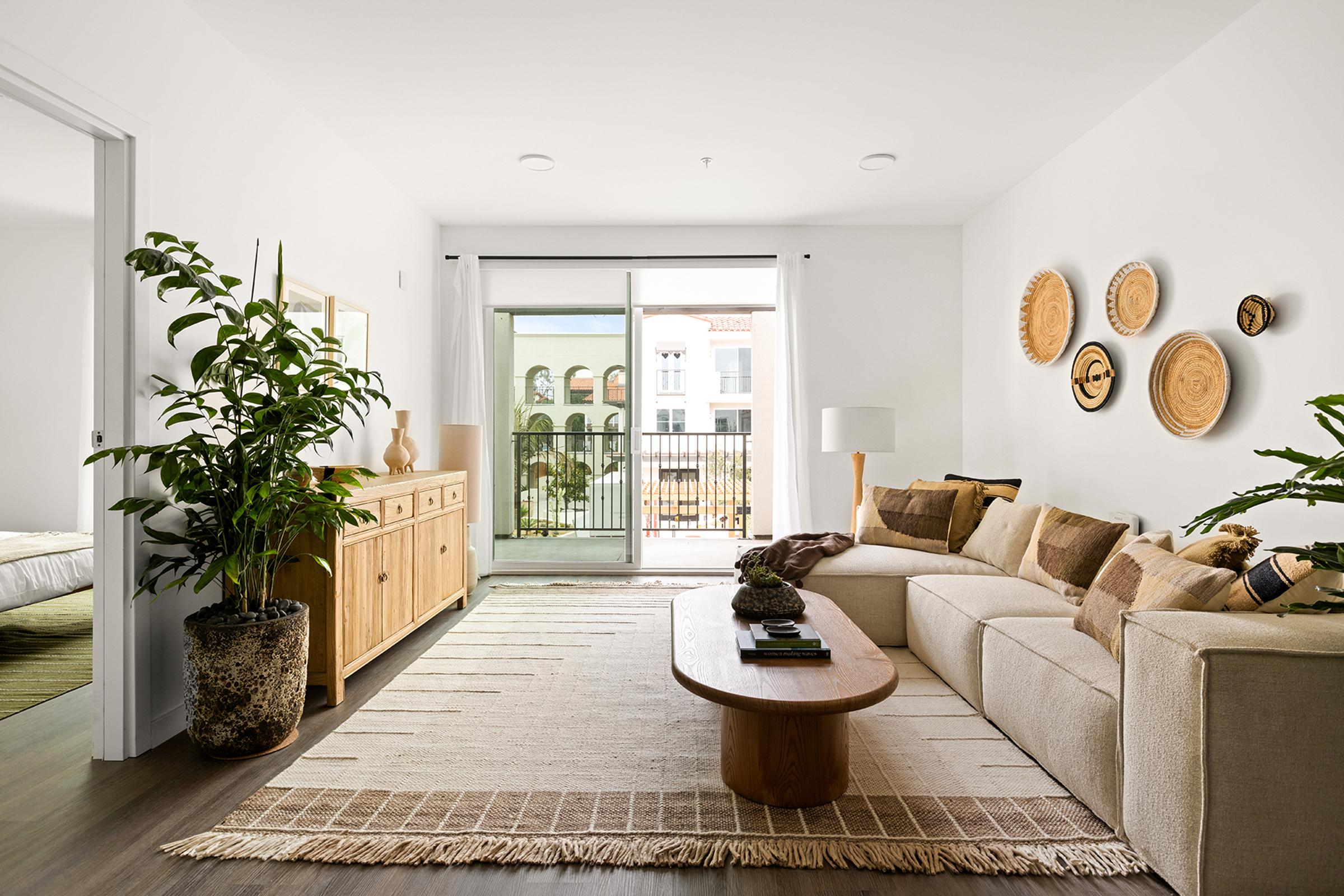 A modern living room featuring a beige sectional sofa adorned with decorative pillows, a wooden coffee table, and a woven area rug. A large plant adds greenery to the space. The room has a sliding glass door that opens to a balcony, with light-colored walls and natural wood accents in the furniture.