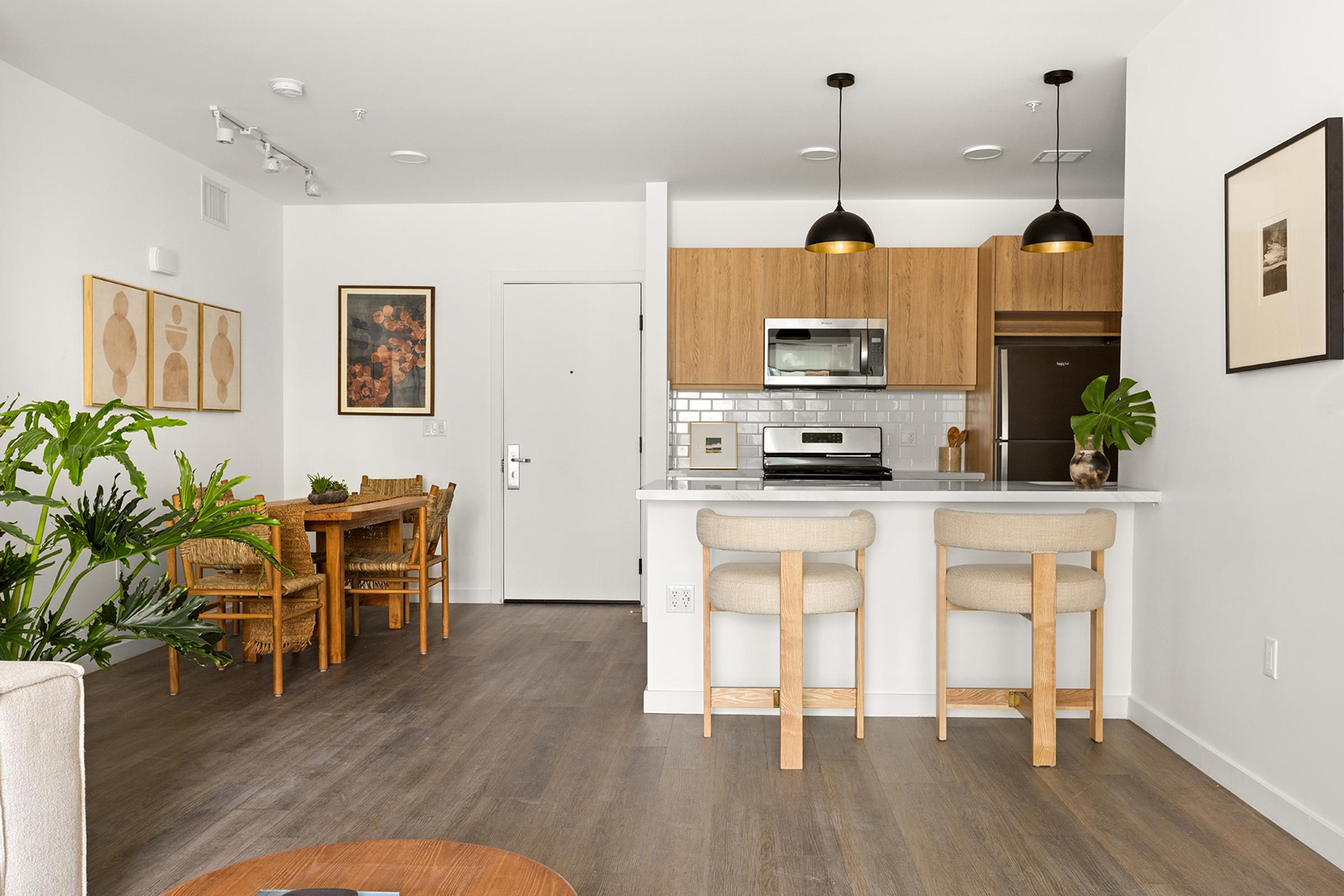 A modern kitchen and dining area featuring light wood cabinets, stainless steel appliances, and a dining table with wicker chairs. There are plants in the room, large windows providing natural light, and minimalistic decor with framed artwork on the walls, creating a warm and inviting atmosphere.