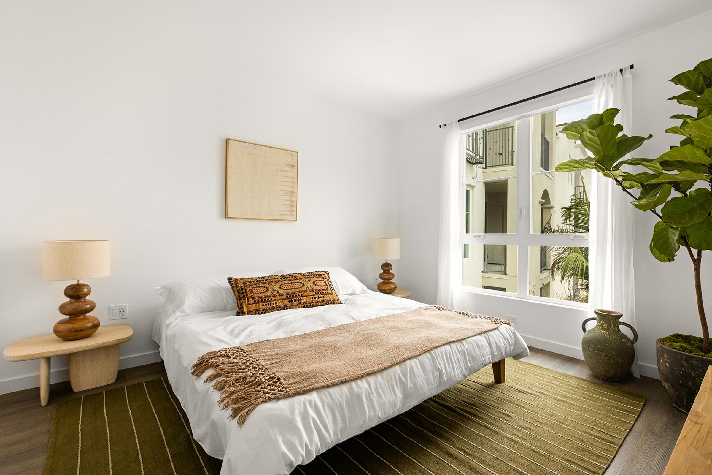 A cozy, modern bedroom featuring a bed with a white duvet, decorative pillows, and a woven throw. There's a large window with sheer curtains allowing natural light, two wooden bedside lamps, and a potted plant, creating a fresh and inviting atmosphere. A decorative rug adds warmth to the floor.