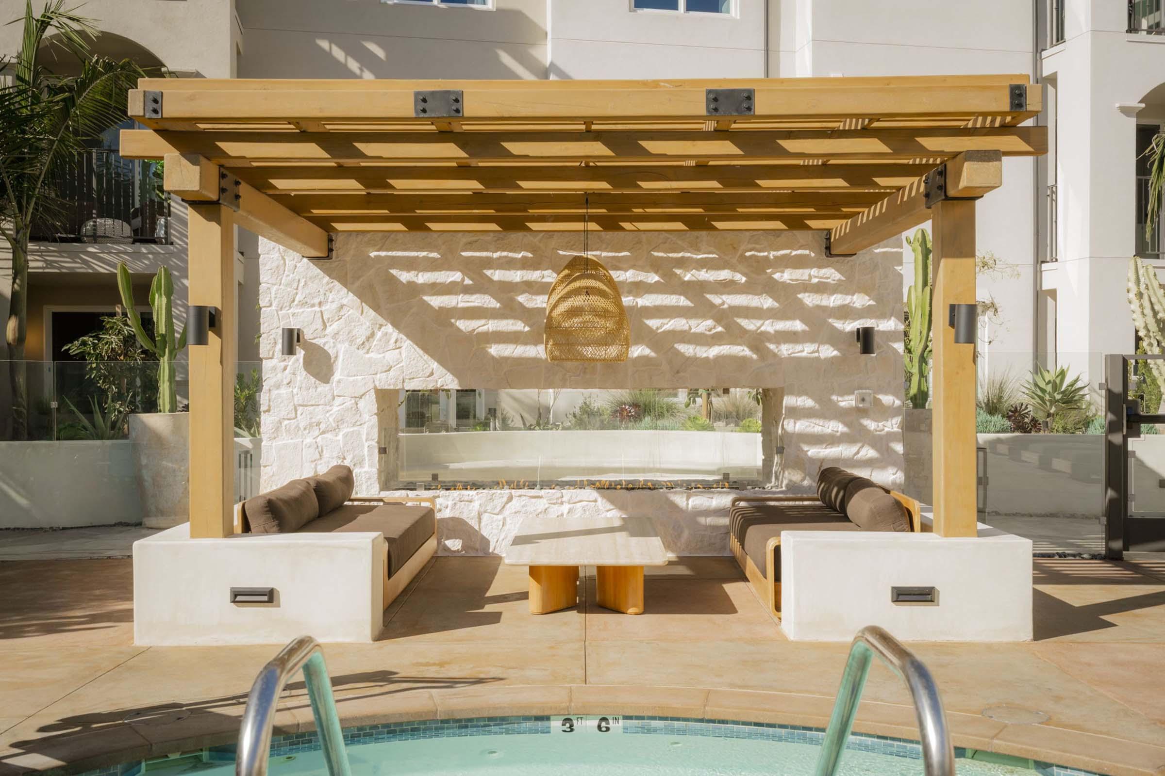 A modern outdoor lounge area near a pool, featuring a wooden pergola with shaded seating on either side. There's a stone wall behind a glass panel, and a small wooden table in the center. Lush greenery and decorative elements can be seen around the area, creating a serene atmosphere.