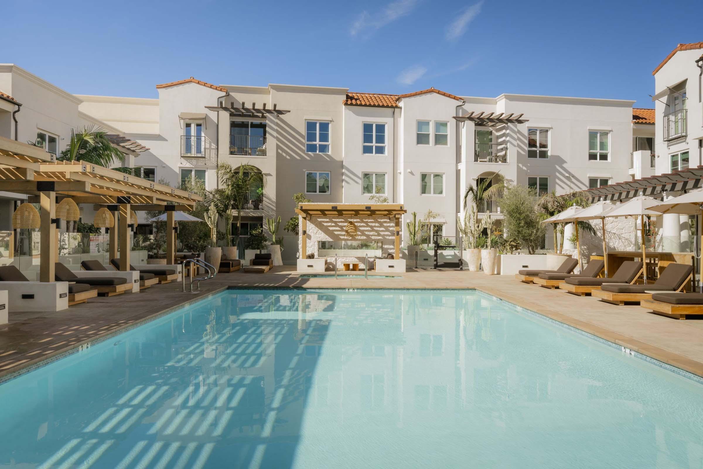 A serene pool area surrounded by modern buildings with a clear blue sky. The pool features lounge chairs and shaded cabanas, creating a relaxing atmosphere. Lush greenery and stylish architectural details enhance the outdoor space, making it ideal for leisure and relaxation.
