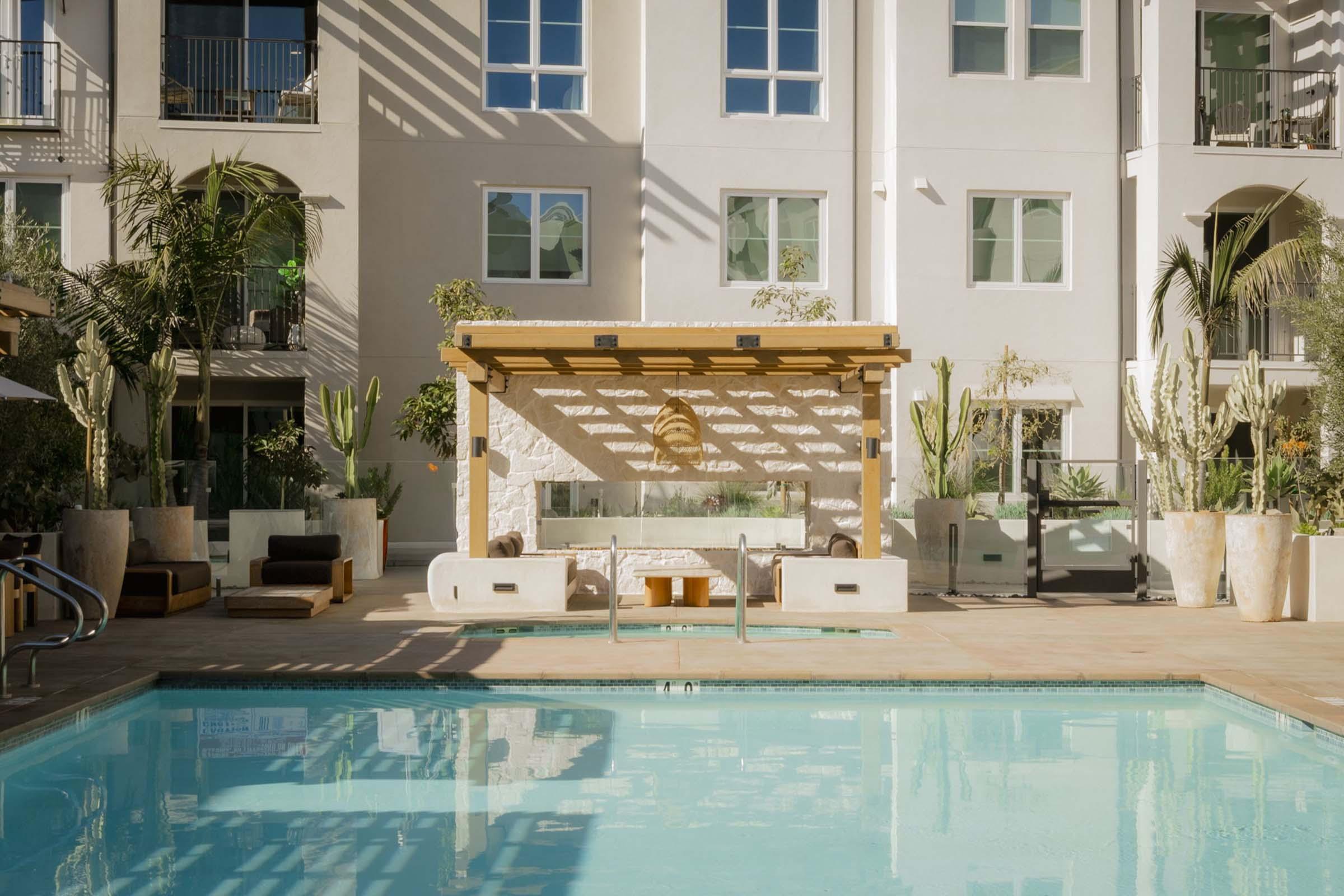 A sunny pool area featuring a clear blue pool in the foreground, surrounded by modern lounge chairs and potted plants. In the background, a stylish outdoor seating area with a wooden pergola, cozy cushions, and decorative cacti, set against a contemporary building with large windows.