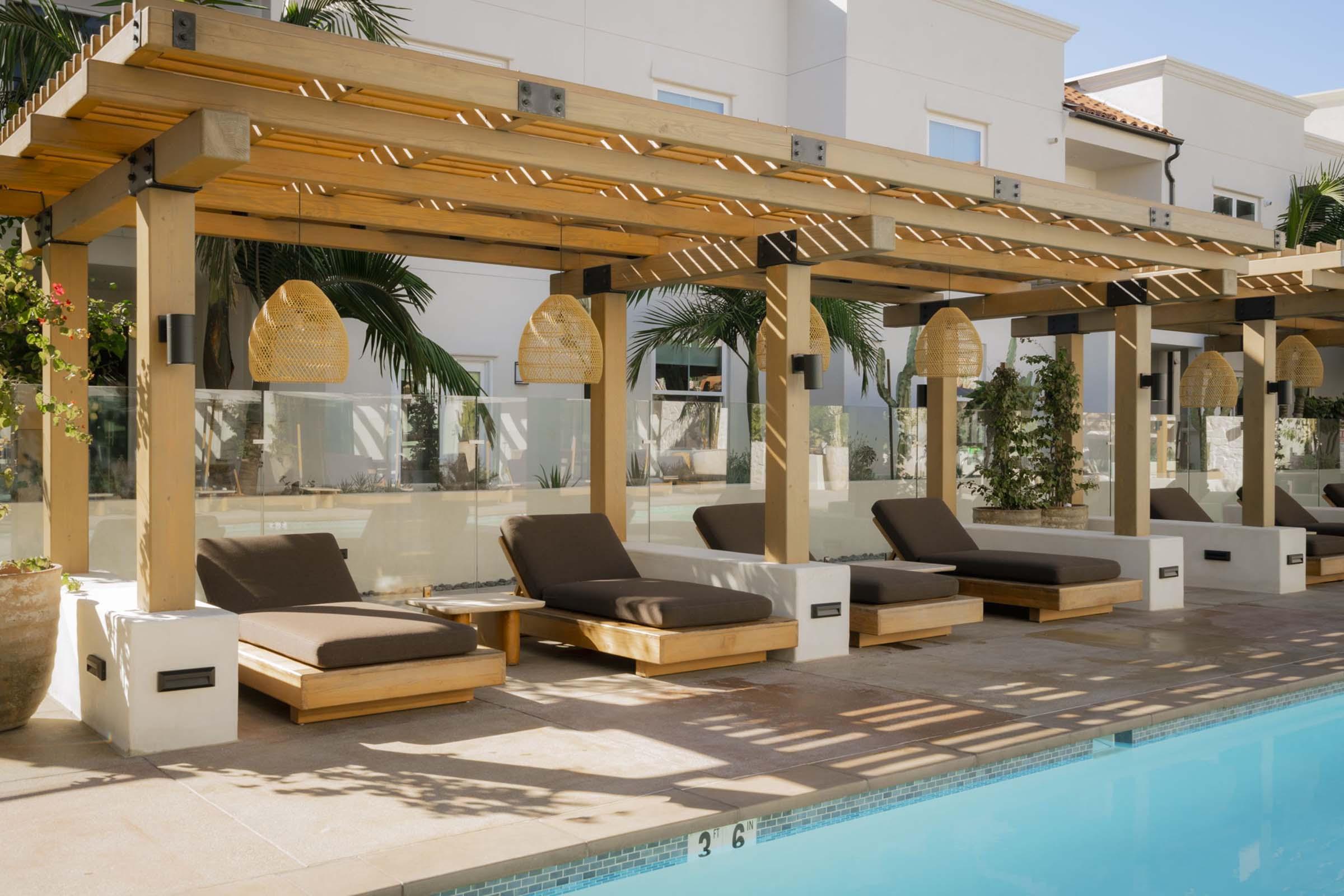 A serene poolside area featuring wooden pergolas with woven lanterns, comfortable brown lounge chairs, and lush greenery in a modern setting. The clear blue water of the pool reflects the sunlight, creating a relaxing atmosphere perfect for leisure.