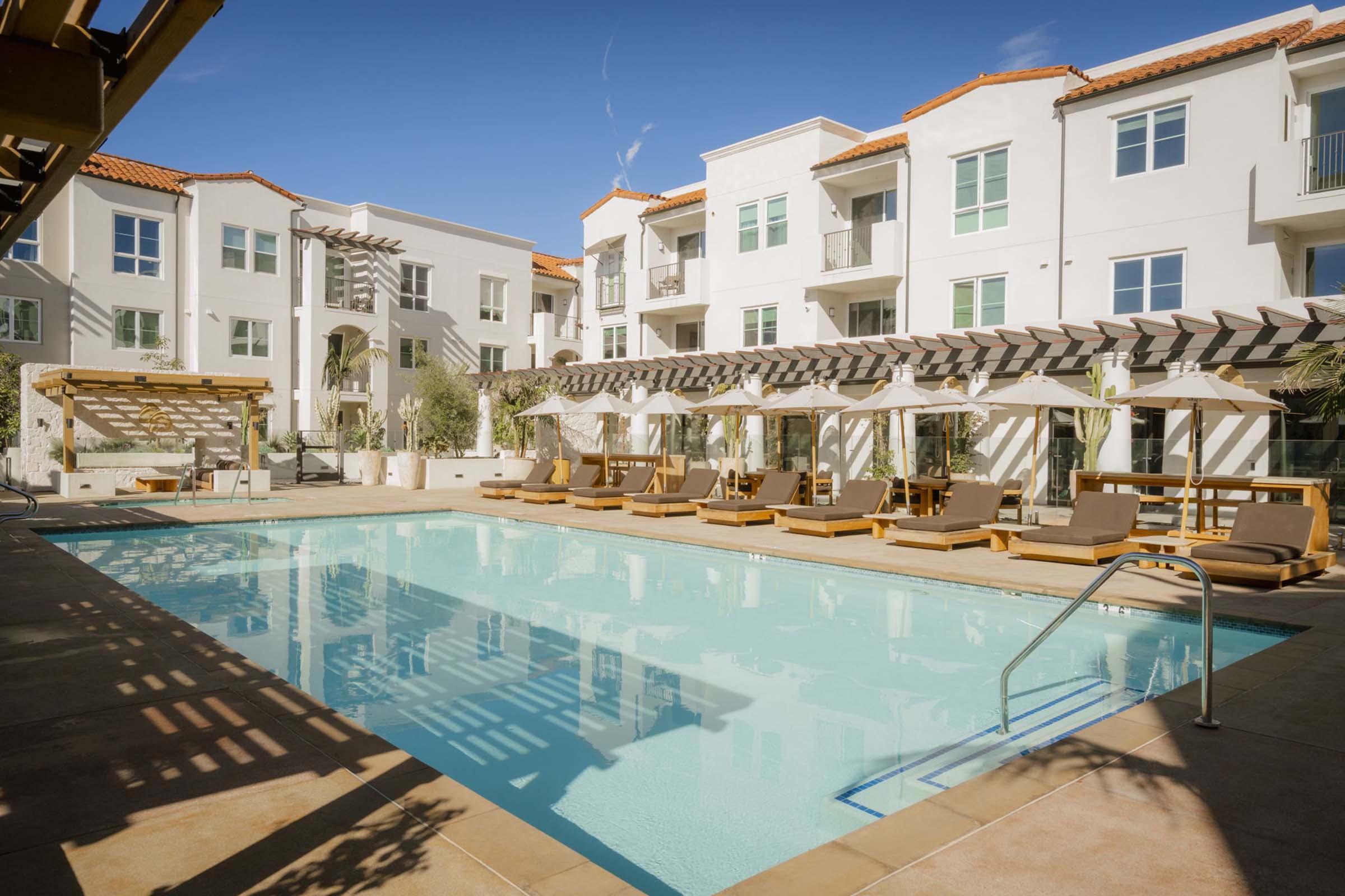 A modern outdoor swimming pool surrounded by lounge chairs and shaded areas, set within a residential complex featuring white buildings with red tile roofs, under a clear blue sky. Sunlight casts shadows from the pergolas onto the pool area.