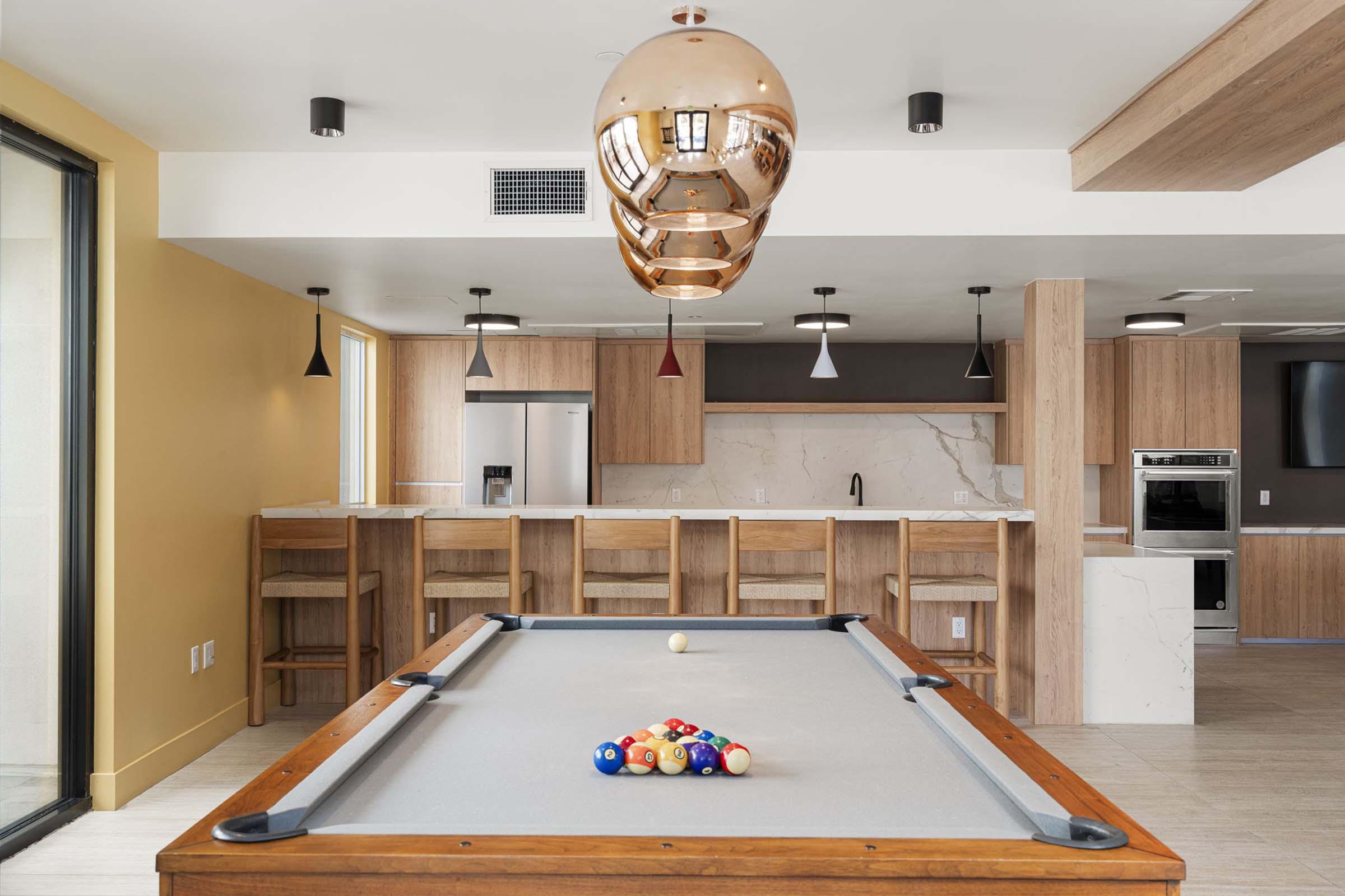 A modern game room featuring a pool table with a triangle rack of balls. In the background, a stylish kitchen with wooden cabinetry, a silver refrigerator, and a marble countertop is visible. The space is well-lit with pendant lighting, and large windows allow for natural light.