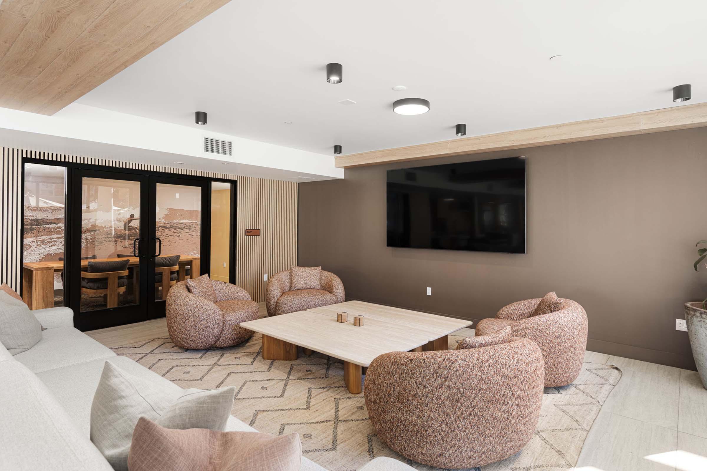 A modern lounge area featuring a cozy arrangement of large, textured bean bag chairs around a low wooden table. The room has warm color tones, a large flat-screen TV mounted on the wall, and a patterned rug beneath the seating. A glass door leads to another space, creating an open and inviting atmosphere.