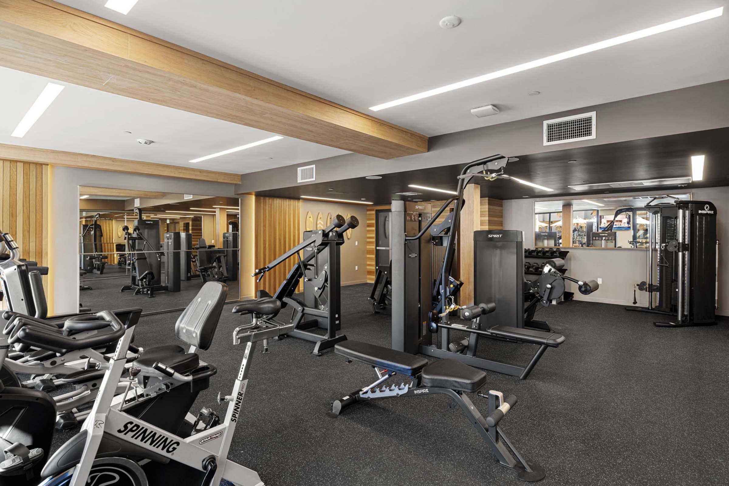 A modern gym interior featuring various exercise equipment, including weight machines, benches, and cardio machines like stationary bikes. The space has wooden accents and large mirrors, providing a bright and inviting atmosphere for workouts.