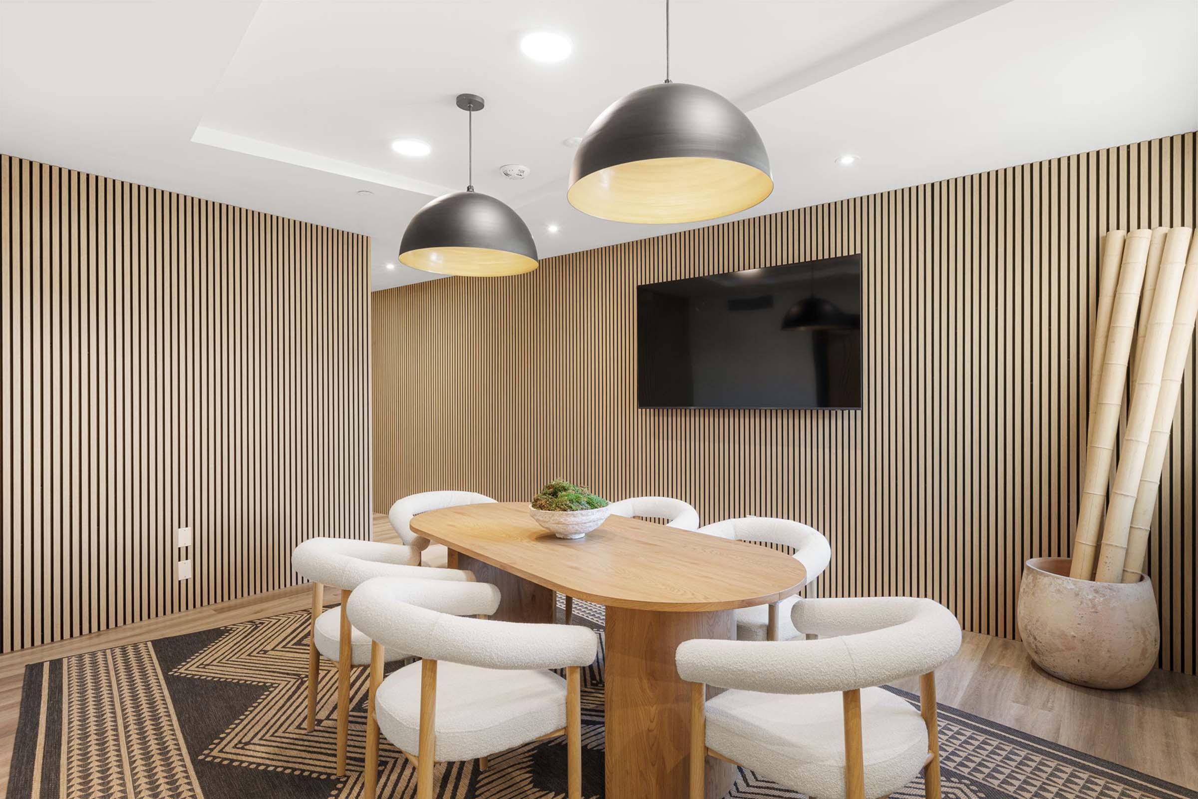 A modern meeting room featuring a round wooden table surrounded by white upholstered chairs. The room has light wood paneling on the walls, two pendant light fixtures hanging from the ceiling, a wall-mounted TV, and decorative elements like a vase and a potted plant in the corner.