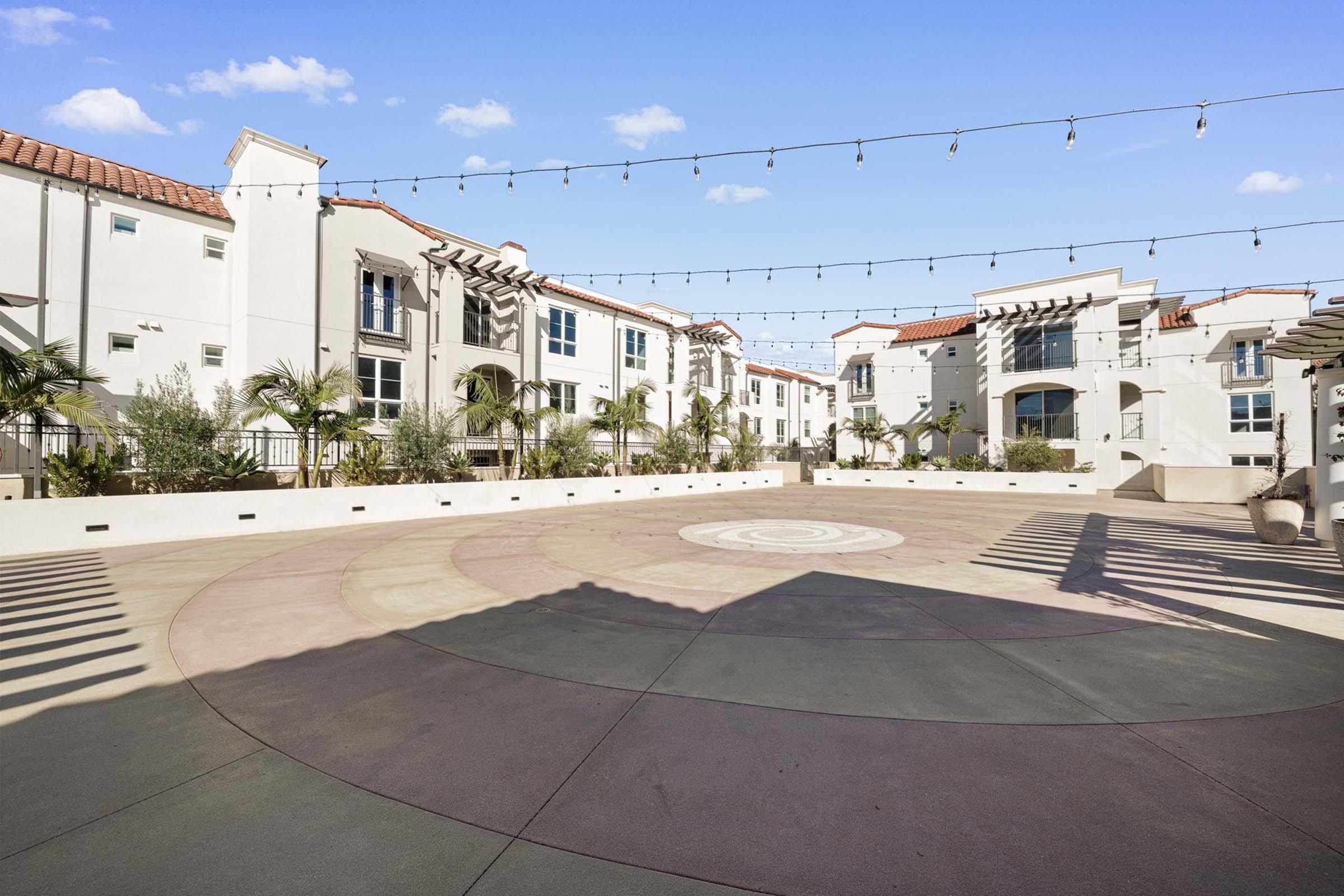 A spacious outdoor courtyard featuring circular patterned flooring, surrounded by modern residential buildings. String lights are hung overhead, and there are palm trees and greenery in the background, creating a welcoming atmosphere for relaxation or gatherings.