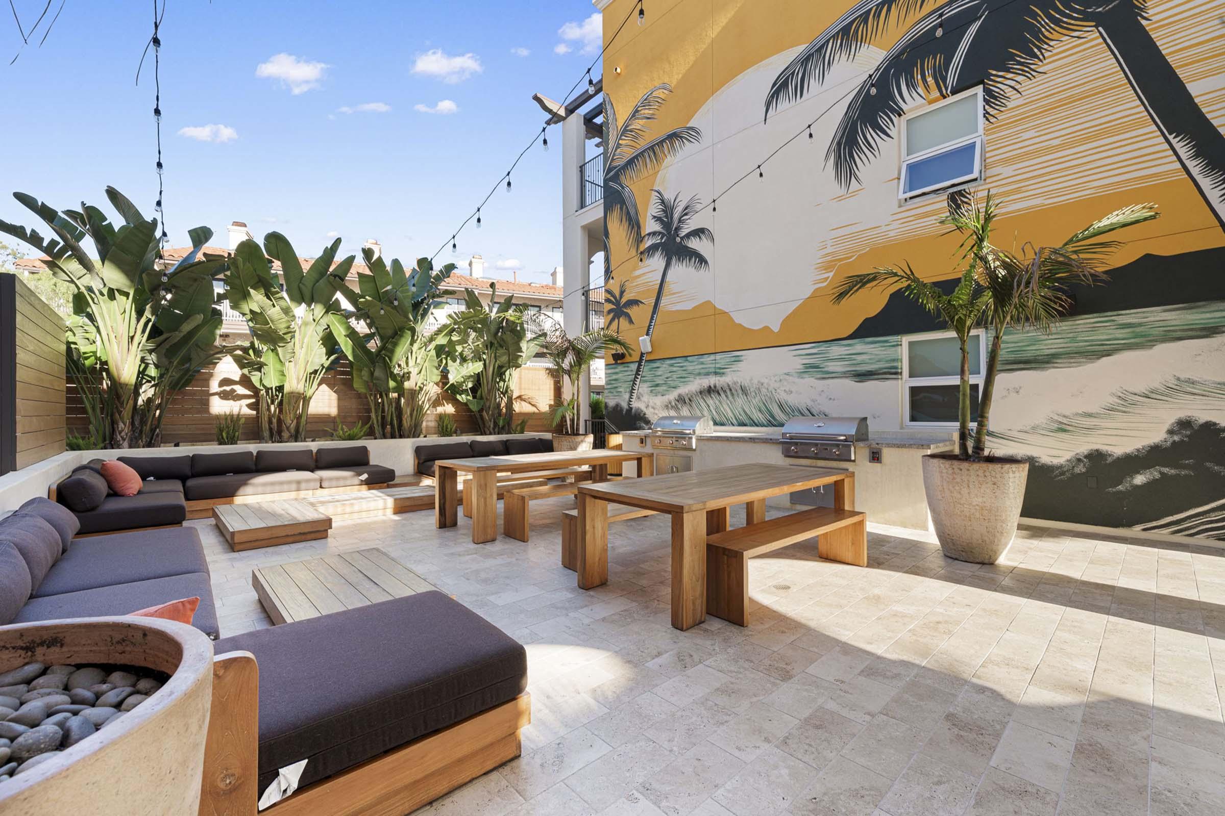 Outdoor lounge area featuring modern wooden furniture, cozy seating with cushions, and BBQ grills. The space is surrounded by lush greenery and a vibrant mural of palm trees and ocean waves on a yellow wall. Strings of lights hang overhead, creating a warm and inviting atmosphere.