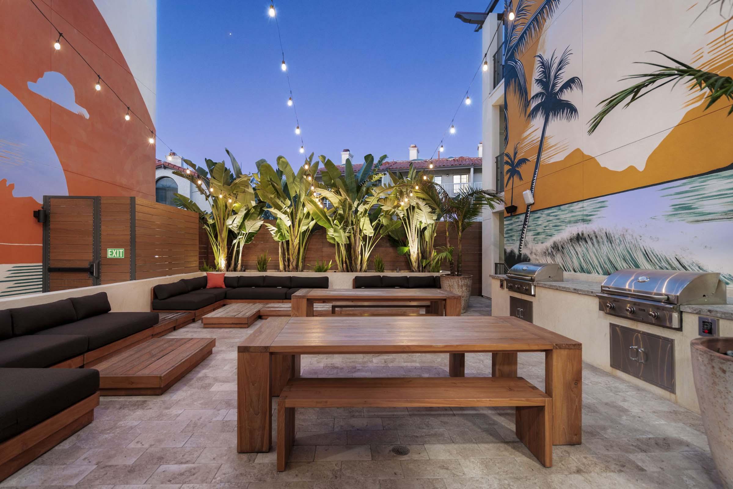 Outdoor patio area featuring wooden furniture, including benches and tables, surrounded by tropical plants and colorful murals on the walls. String lights hang above, creating a cozy atmosphere at dusk. Barbecue grills are set up for outdoor cooking.