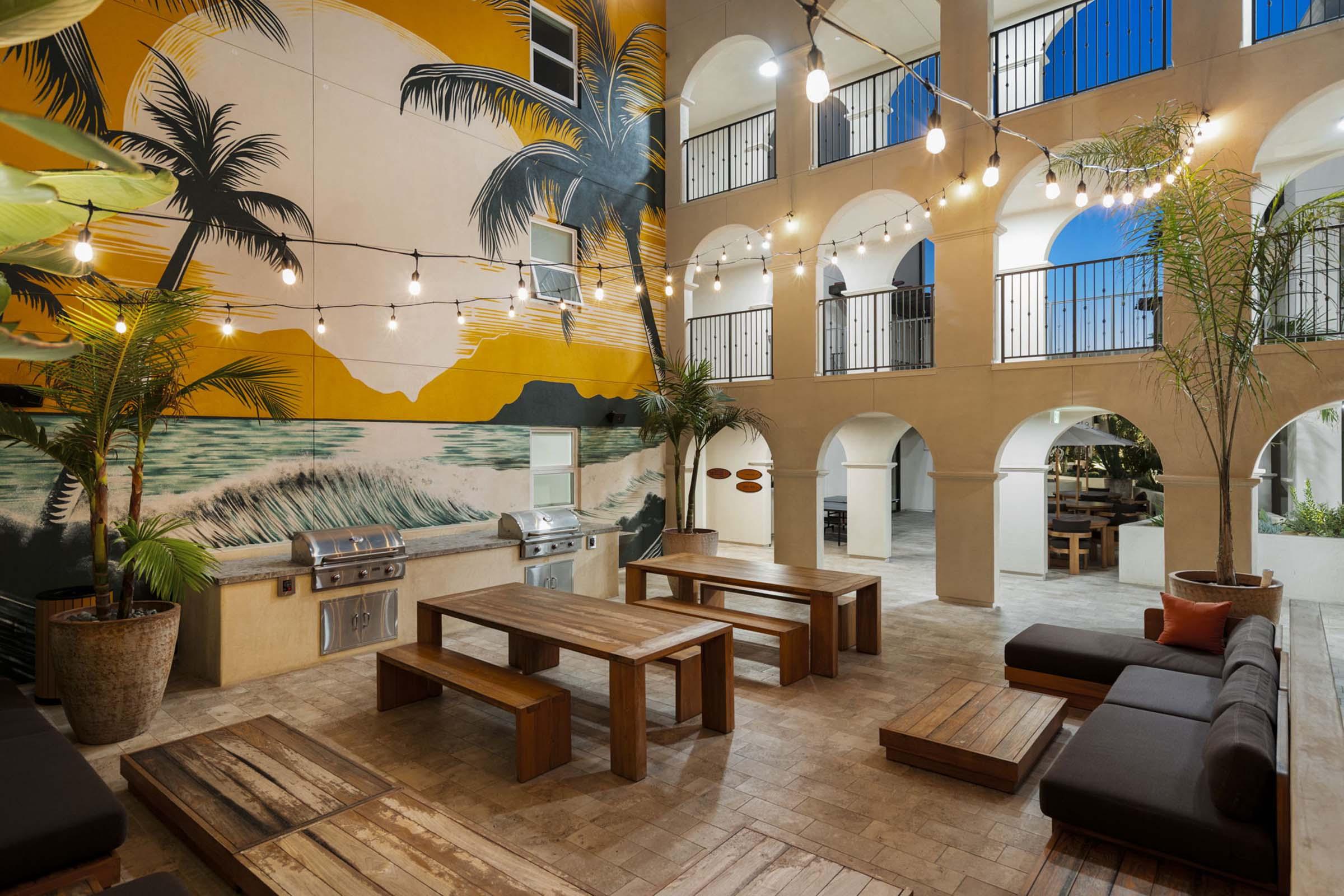 An outdoor courtyard featuring vibrant palm tree murals on the walls, string lights overhead, and multiple seating areas with wooden and cushioned furniture. The space includes barbecue grills and is surrounded by arches, creating a relaxing atmosphere.
