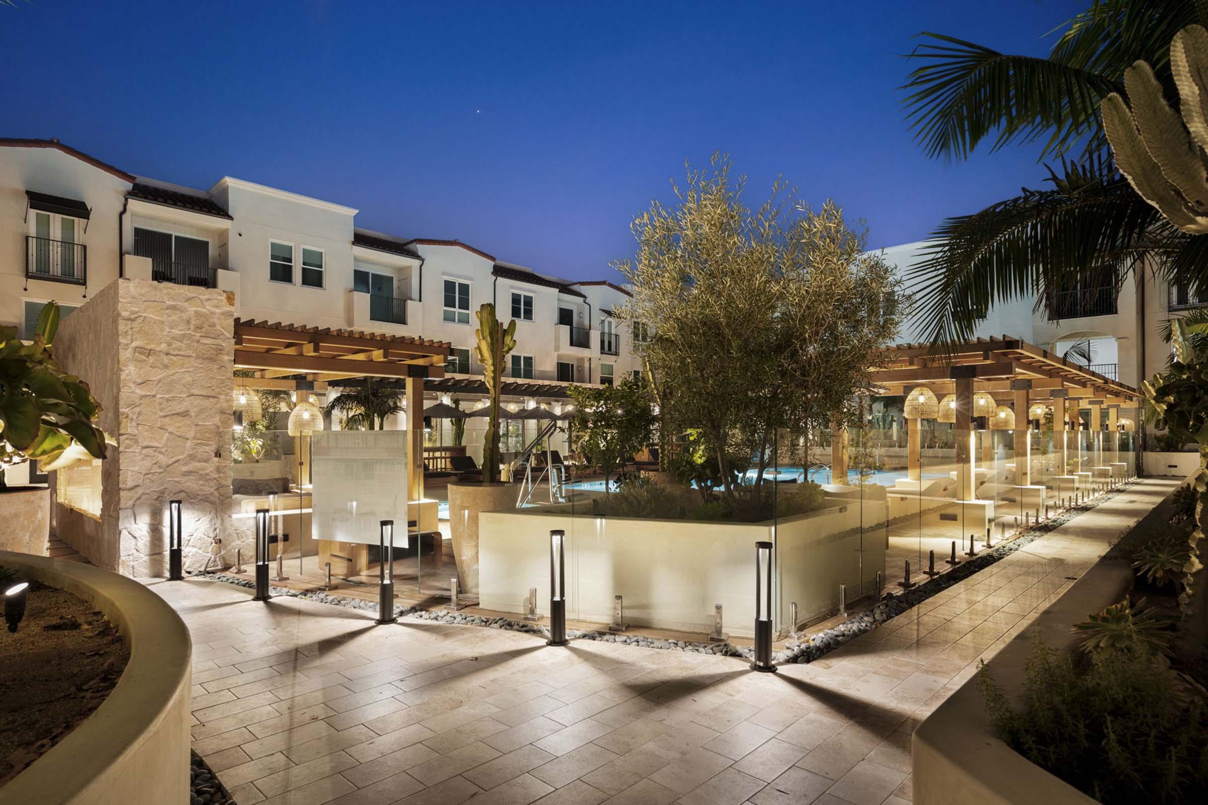 A well-lit outdoor area of a modern resort at dusk, featuring landscaped gardens, a swimming pool with lounge chairs, and stylish outdoor furniture. The architecture includes multiple buildings with balconies, surrounded by palm trees and illuminated pathways.