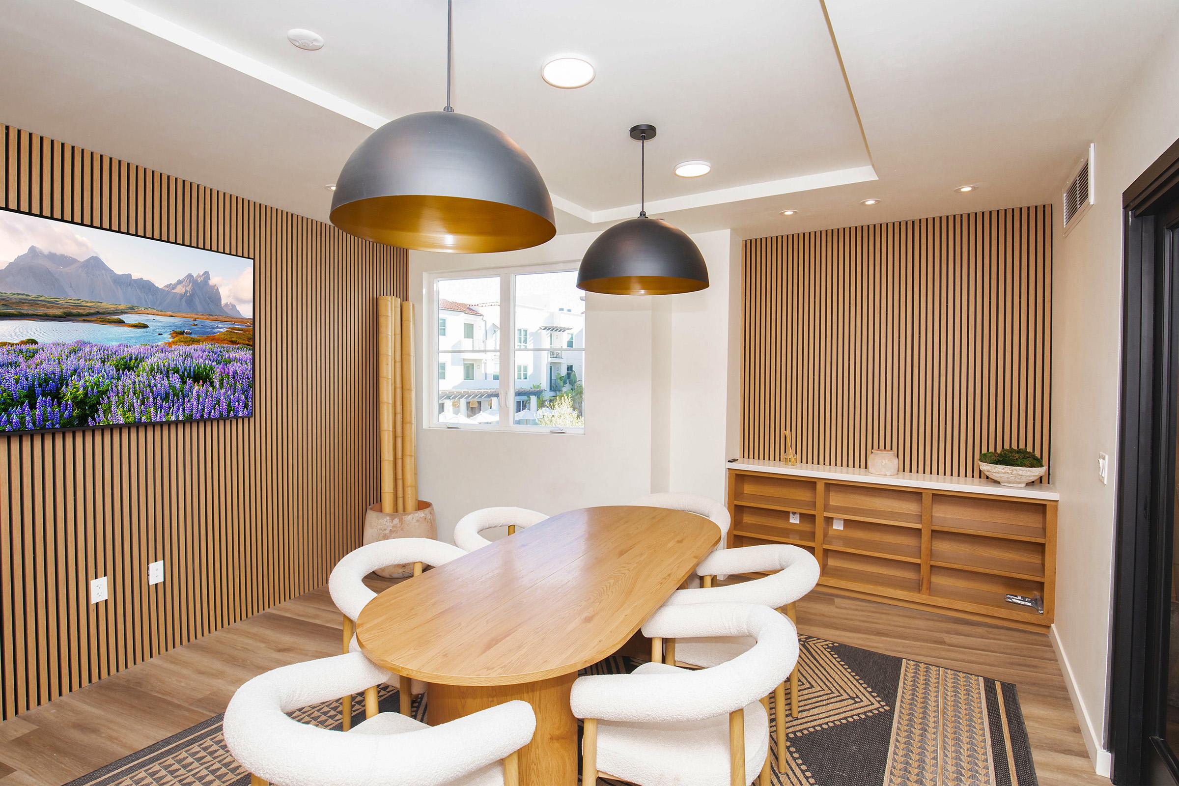 A modern meeting room featuring a large oval wooden table surrounded by upholstered chairs. Two stylish pendant lights hang from the ceiling. A wall-mounted television displays a scenic landscape, while the room is decorated with wooden paneling and a minimalist sideboard. Natural light enters through a window.