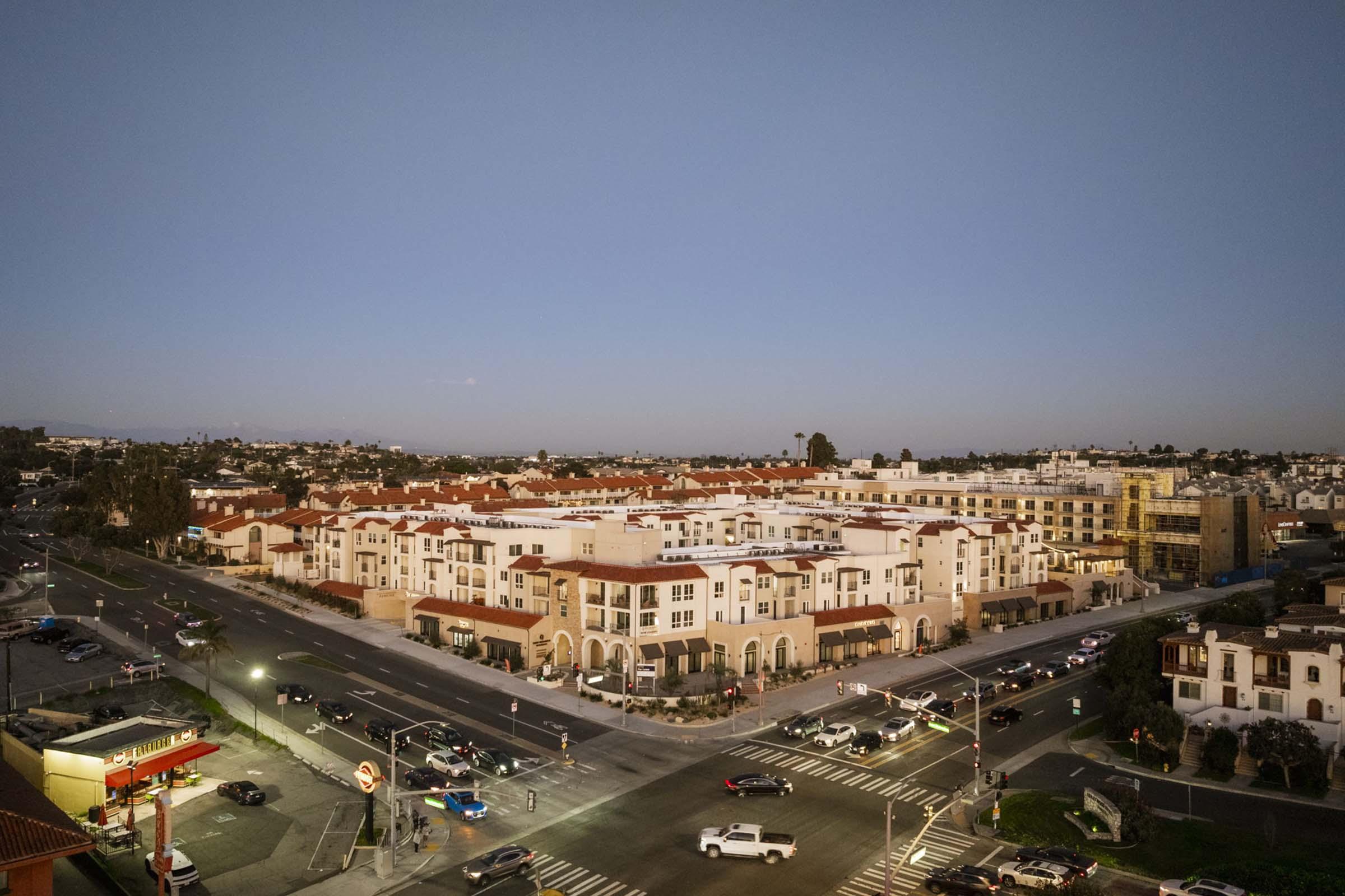 Aerial view of a suburban neighborhood at dusk, showcasing a mix of residential buildings with red-tiled roofs, streets filled with parked cars, and commercial areas in the foreground. Soft evening light casts a serene glow over the scene, highlighting the area's layout and architecture.