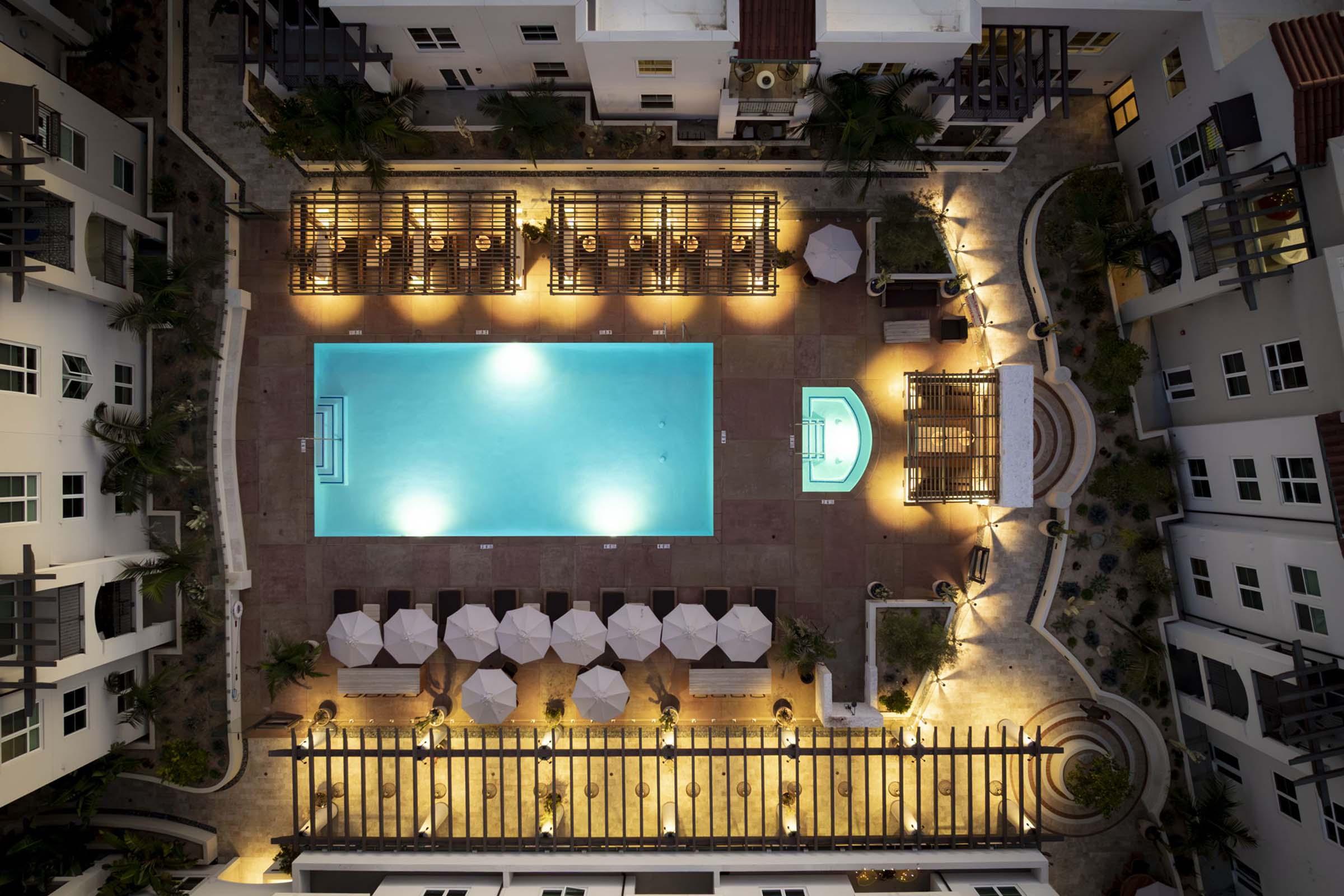 Aerial view of a luxury hotel pool area featuring a large, illuminated swimming pool surrounded by lounge chairs, umbrellas, and wooden pergolas at night. The surrounding buildings are modern, and the atmosphere is serene and inviting, enhanced by warm lighting and landscaped greenery.