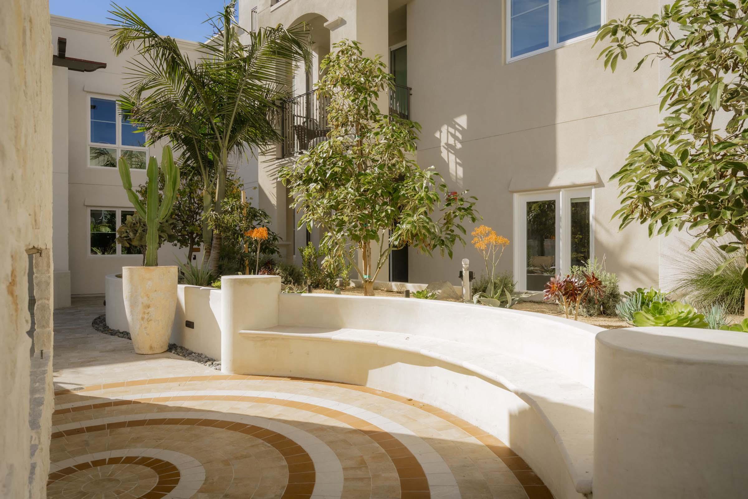 A sunny courtyard featuring a curved white bench surrounded by vibrant greenery, including palm and cactus plants. The space is enhanced with colorful flowers and has a circular patterned stone floor. The walls are light-colored, complementing the bright, open atmosphere.