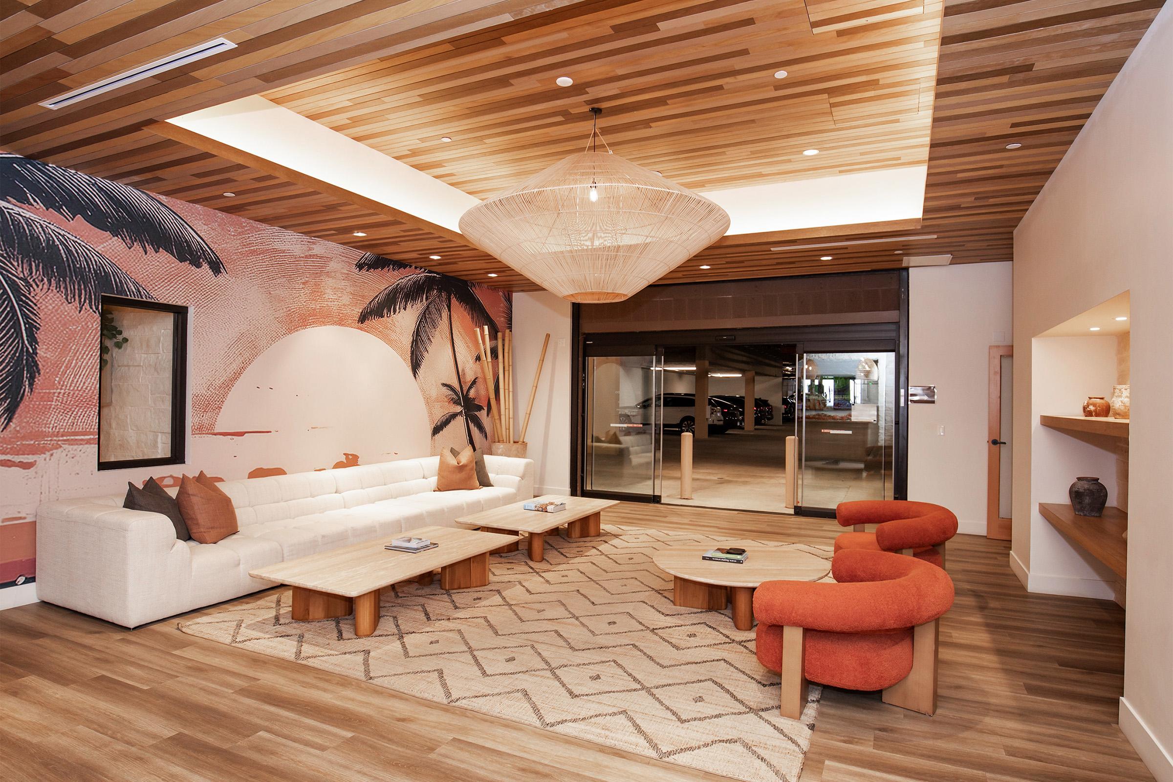 A modern lounge area featuring a white sectional sofa, two orange accent chairs, and a wooden coffee table. The room has a warm wood ceiling and a large wall mural depicting tropical palm trees. Natural light fills the space through a large glass door leading outside. A patterned rug completes the cozy atmosphere.