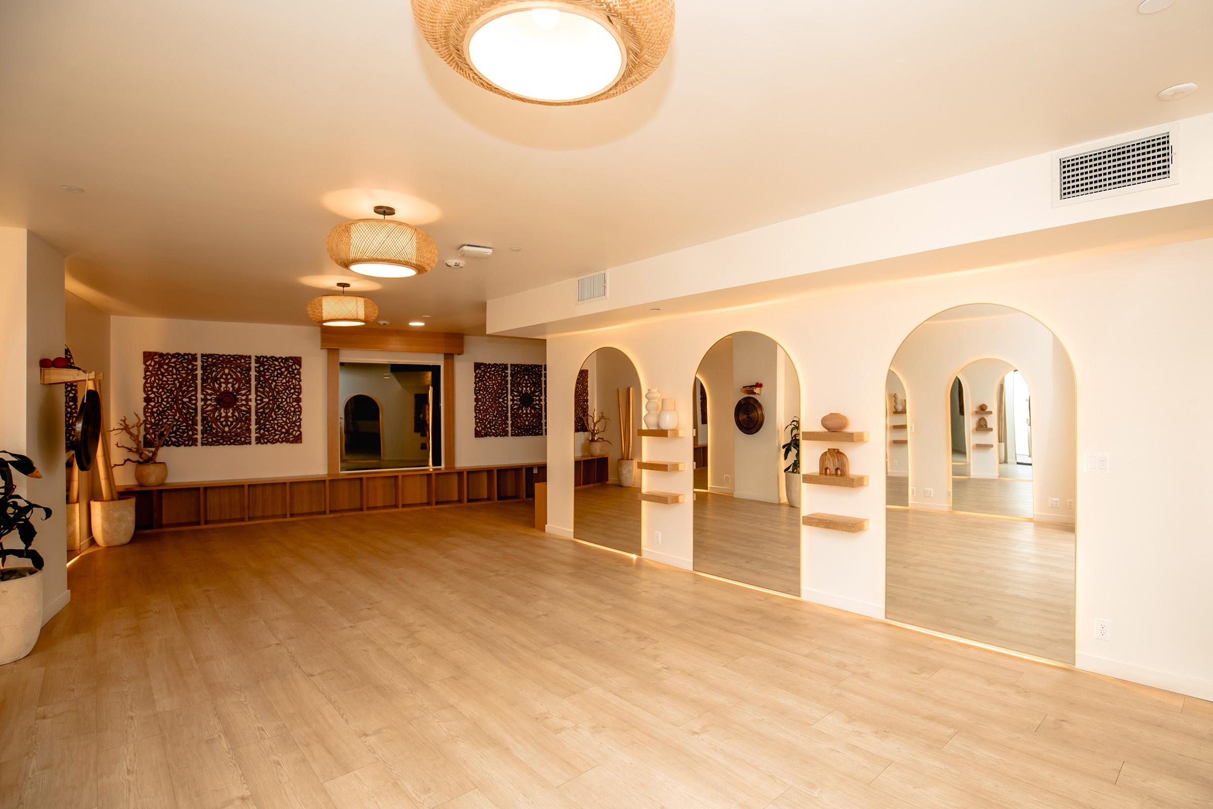 A spacious, well-lit interior featuring wooden flooring, arched wall alcoves with decor, and soft lighting from hanging fixtures. Neutral tones create a calming atmosphere, complemented by minimalistic decorations and potted plants. The area appears open and inviting, suitable for gatherings or wellness activities.