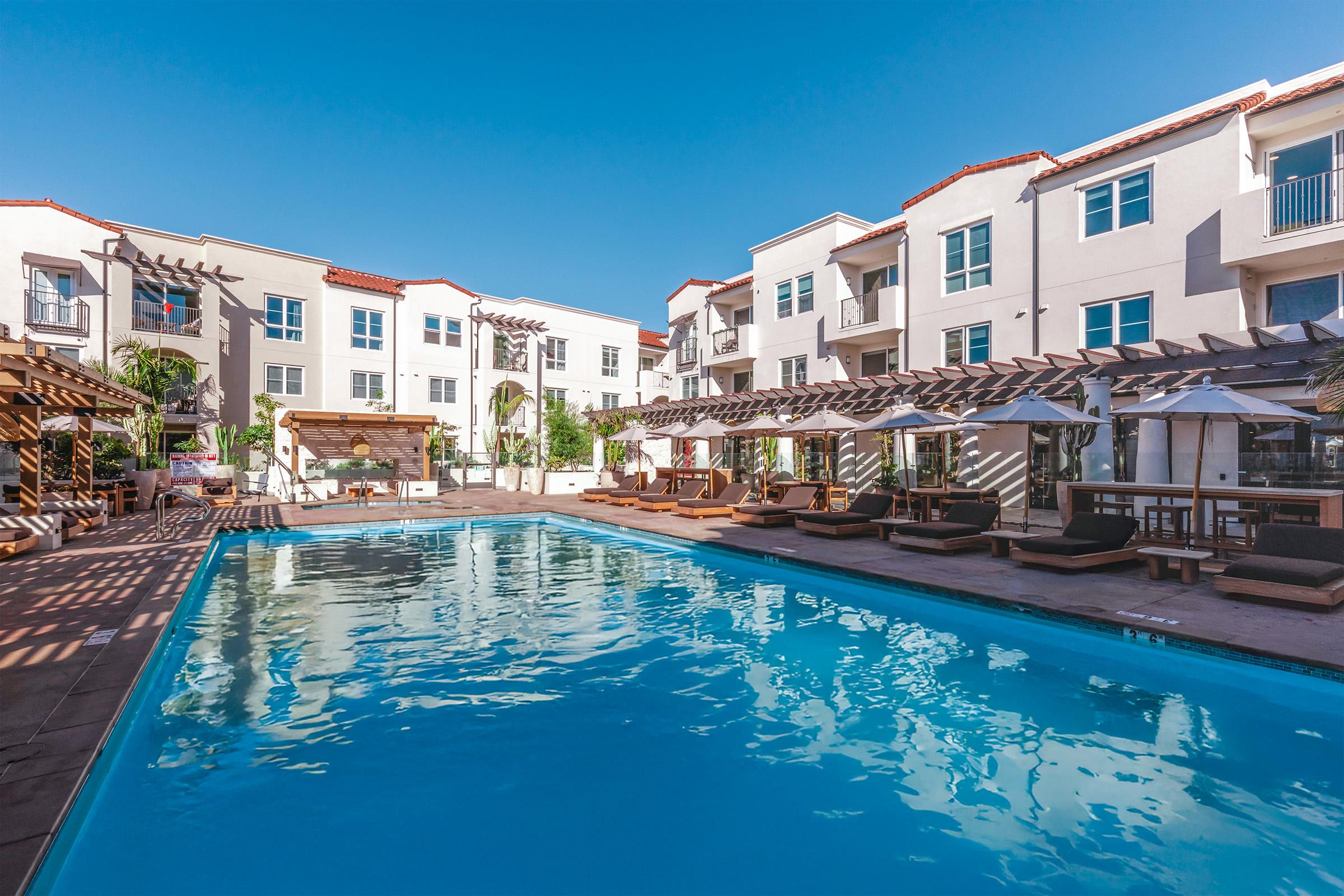 A well-lit, modern apartment complex featuring a large swimming pool surrounded by lounge chairs. In the background, there are multiple buildings with balconies, palm trees, and landscaping. The scene is inviting and captures a sunny day by the poolside.