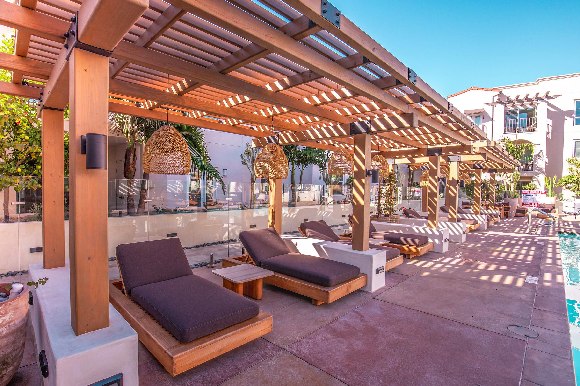 View of a modern outdoor pool area featuring wooden lounge chairs and cabanas under a pergola. Sunlight filters through the slats, casting shadows on the concrete deck. Lush greenery and a serene atmosphere create a relaxing environment suitable for leisure and sunbathing.