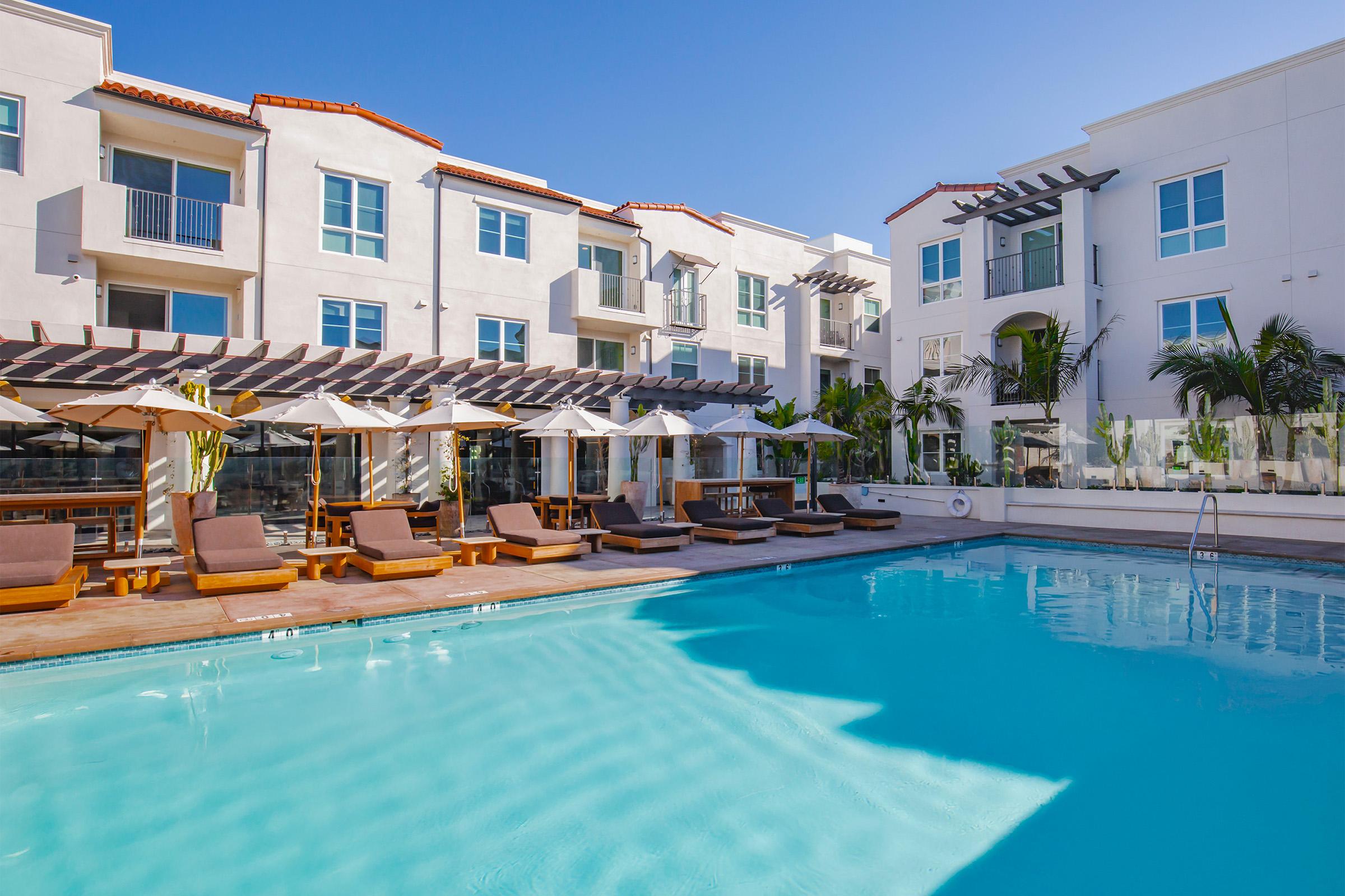 A serene outdoor pool area surrounded by lounge chairs and umbrellas, with modern buildings in the background. The clear blue sky complements the inviting water, creating a relaxing atmosphere for guests to enjoy.